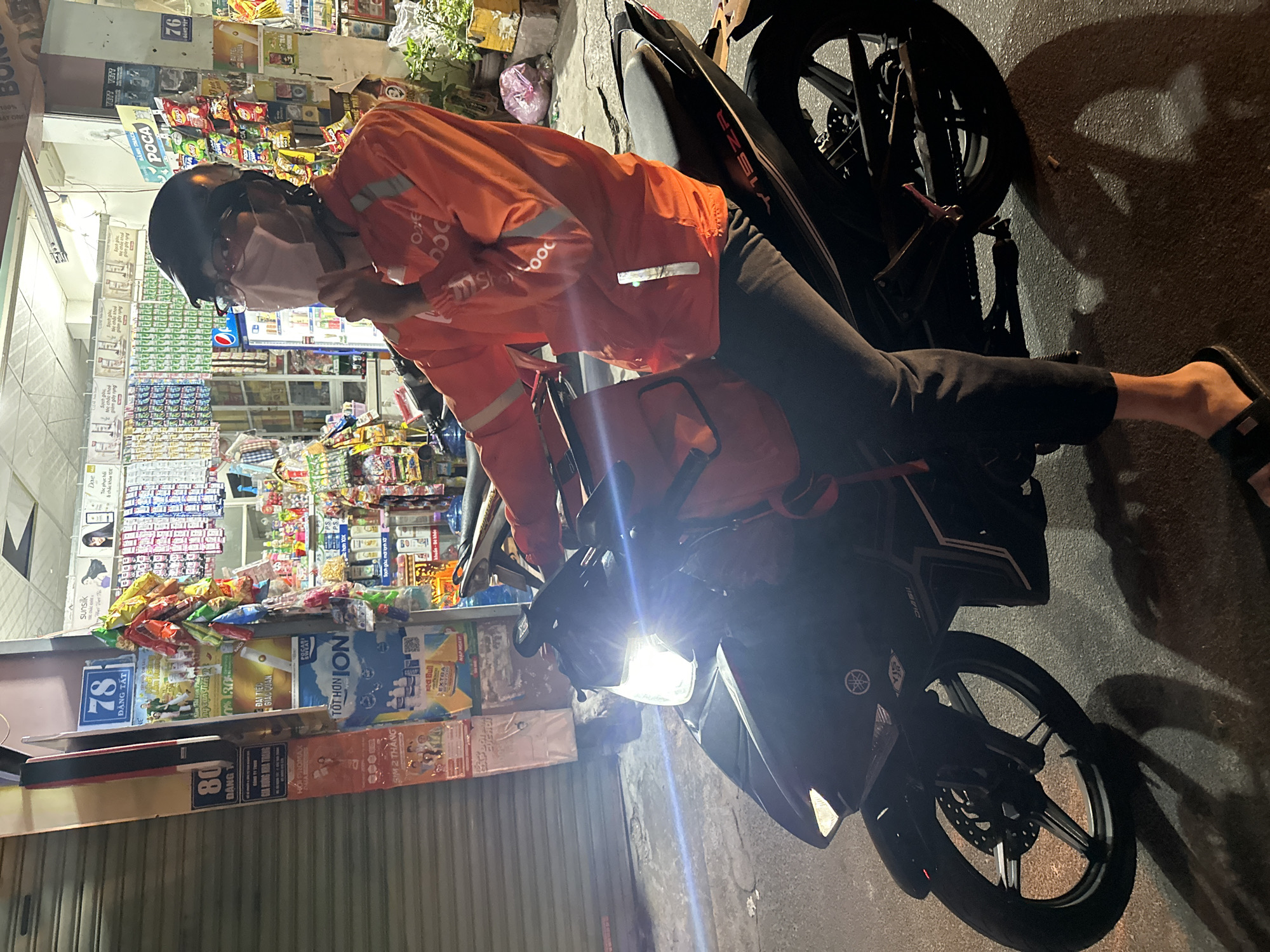 The image shows a person in an orange ShopeeFood jacket sitting on a Yamaha motorcycle outside a convenience store in the city of Bac Nha Trang Ward, Vietnam. The person is wearing a helmet and a face mask. The convenience store displays various snacks and drinks, and advertisements for products like "Red Bull Extra," "Poca" chips, and "Sunsilk" hair products. Signs for "78 Dang Tat" and "80 Dang" are visible. The motorcycle's headlight is on. The image was taken at night.
