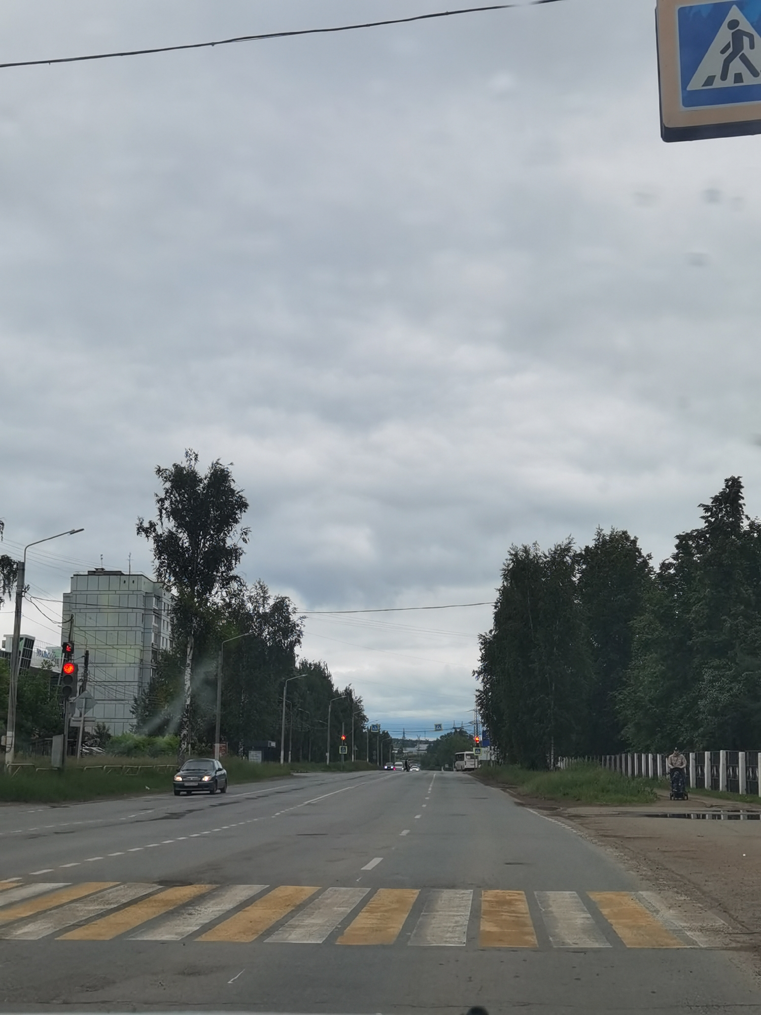 The image presents a street scene in Chaikovsky, Russia, observed from a vehicle on an overcast day. A multi-lane asphalt road extends into the distance, marked by a pedestrian crosswalk in the foreground featuring alternating yellow and white stripes.
On the left side of the road, a dark-colored sedan is stationary at an intersection, where a red traffic light is illuminated on a pole. Behind the pole and surrounding foliage, a multi-story building with a light-colored, gridded facade is visible.
On the right side of the road, a person in light clothing is walking on a dirt path, pushing a dark-colored stroller. This path is bordered by several puddles and runs parallel to a fence constructed from alternating dark and light vertical segments. A dense line of trees lines the roadside, extending into the background.
In the upper right corner, a blue and white rectangular pedestrian crossing sign is partially visible. A dark power line stretches horizontally across the top of the frame. The sky is uniformly gray and overcast, indicating daytime. Additional vehicles and another red traffic light are discernible further down the road in the distance.