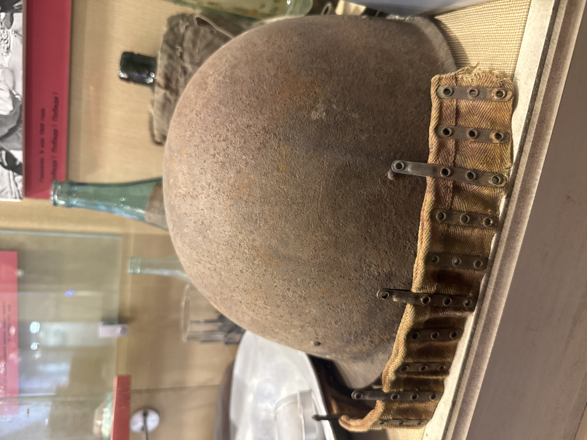 The image depicts a rusty, metallic helmet with a fabric strap, likely a military artifact. It is displayed in a glass case, suggestive of a museum setting. The background includes various objects, such as bottles, and a book with text in Cyrillic script. The context of these background objects indicates the location is likely a museum or historical display in Saint Petersburg, Russia.