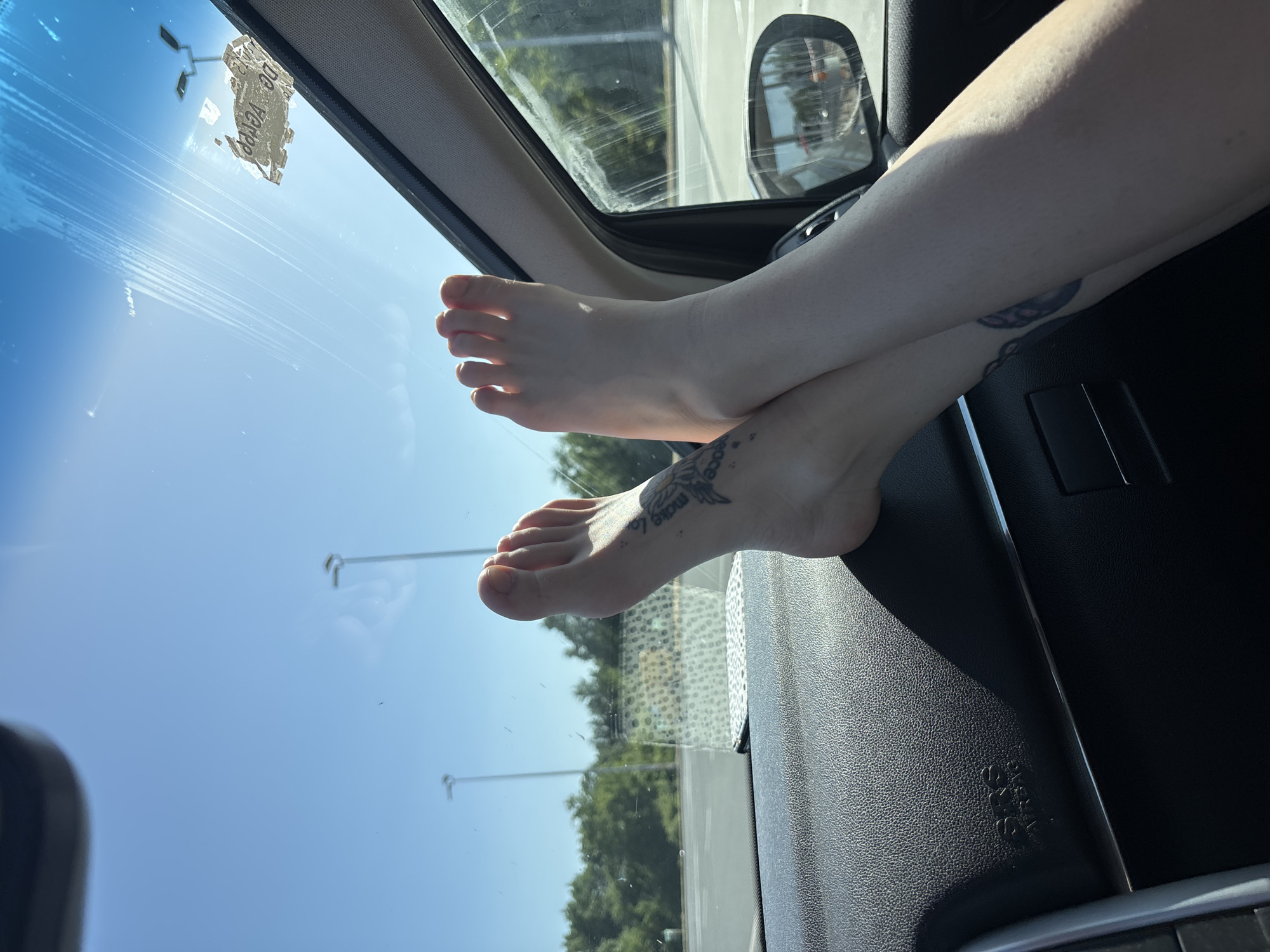 The image depicts two bare feet resting on the dashboard of a car. The feet have tattoos on them. The car appears to be parked, and through the windshield, one can see streetlights and trees. The location is identified as Obrenovac Urban Municipality, Serbia, based on the reflection on the windshield.