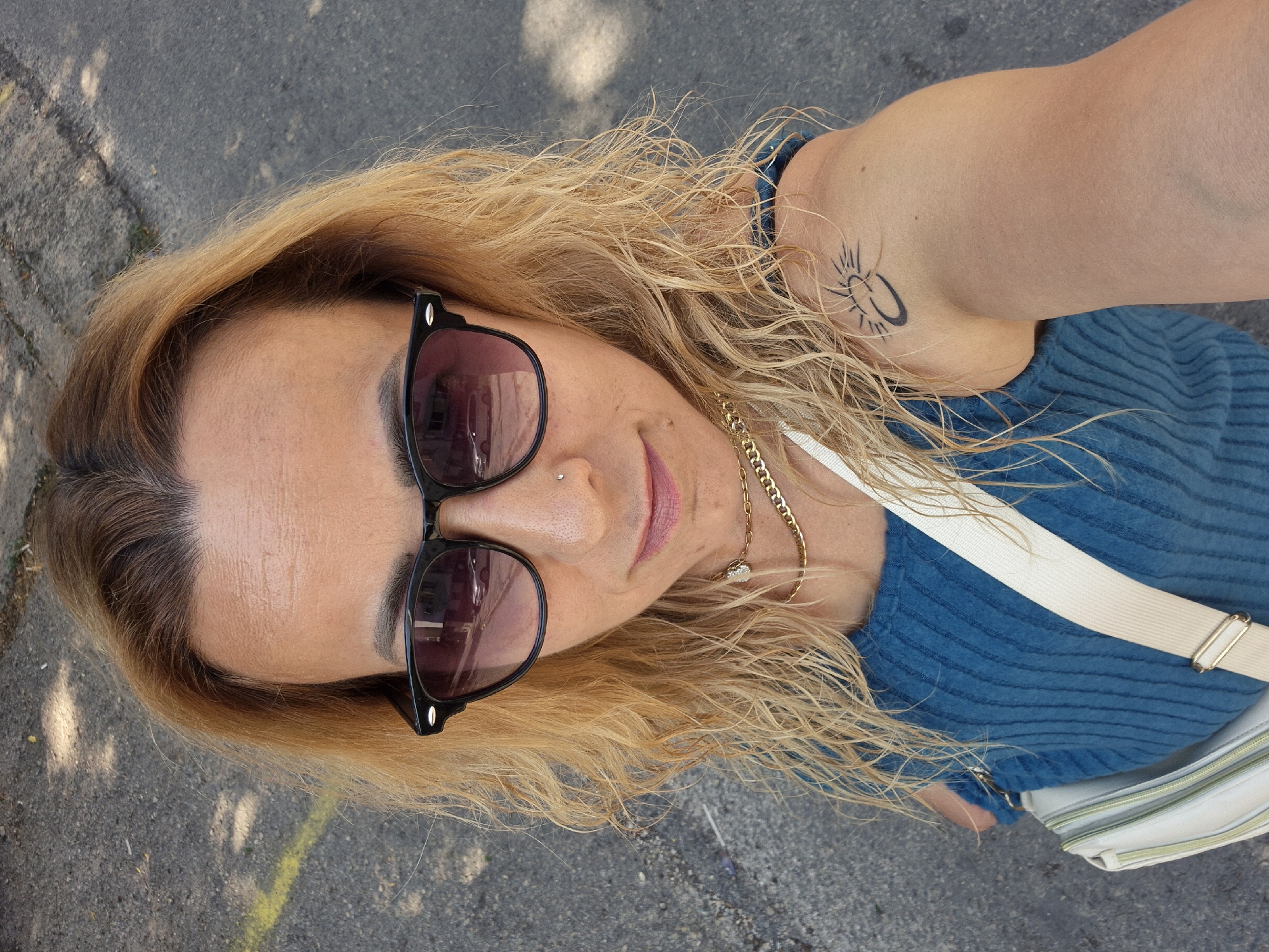 A woman is taking a selfie outdoors. She has blonde, wavy hair and is wearing dark sunglasses, a blue ribbed top, and a gold necklace. She also has a small nose ring and a tattoo of a sun on her right shoulder. A light-colored crossbody bag is slung across her chest. The background appears to be a paved surface, possibly a street or sidewalk, with some dappled sunlight and shadows. There is a yellow line painted on the ground to the left of the woman. The city and specific location are unknown.