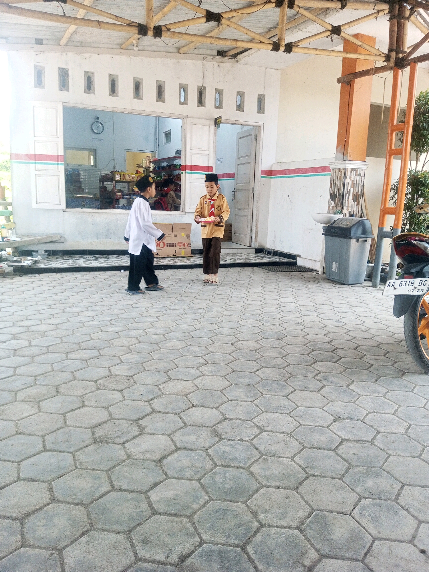 Two young boys, dressed in traditional Islamic attire including peci hats, stand on a hexagonal paved courtyard. One boy, wearing a white tunic and black pants, walks towards the right. The other boy, in a tan uniform with a red scarf, holds a plate of food. They are positioned in front of a building with a white facade, red and green striped trim, and open windows. A motorcycle is partially visible on the right side of the image, with its license plate reading "AA 6319 BG." The scene appears to be outdoors, possibly in front of a school or religious institution in the city of Kalibeber, Indonesia.