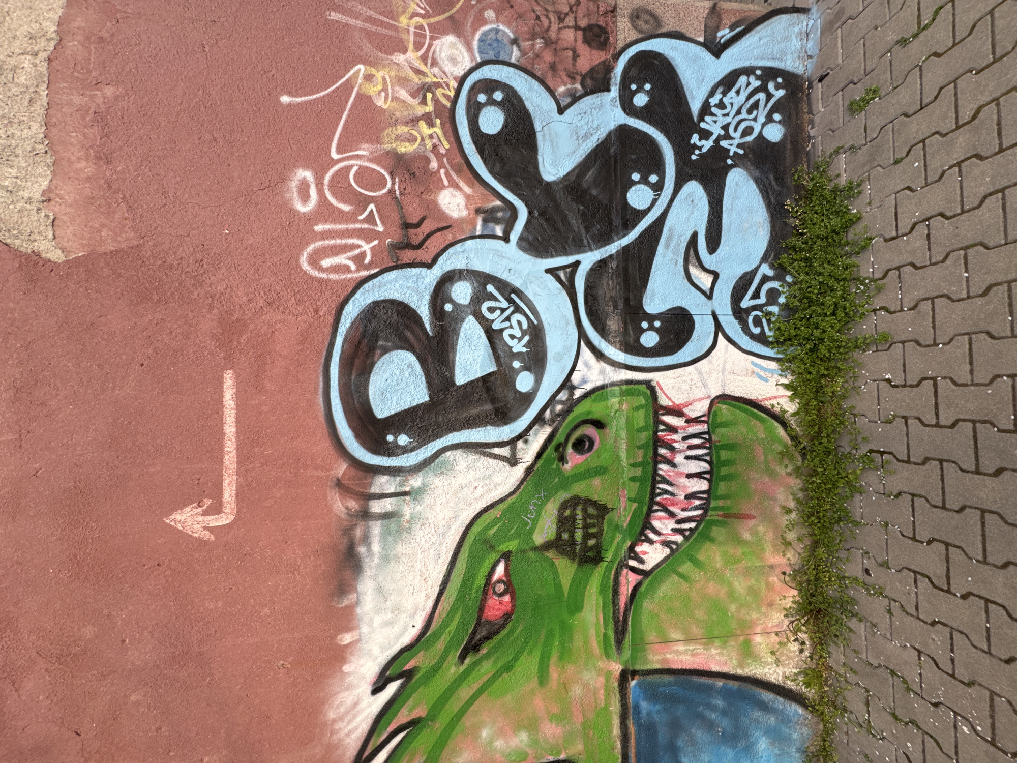 The image captures graffiti on a brick wall in Sofia, Bulgaria. The graffiti includes bubble letters in blue and black, with a depiction of a green dinosaur-like creature with sharp teeth. An arrow points towards the graffiti. Ivy grows along the edge of the brick wall.