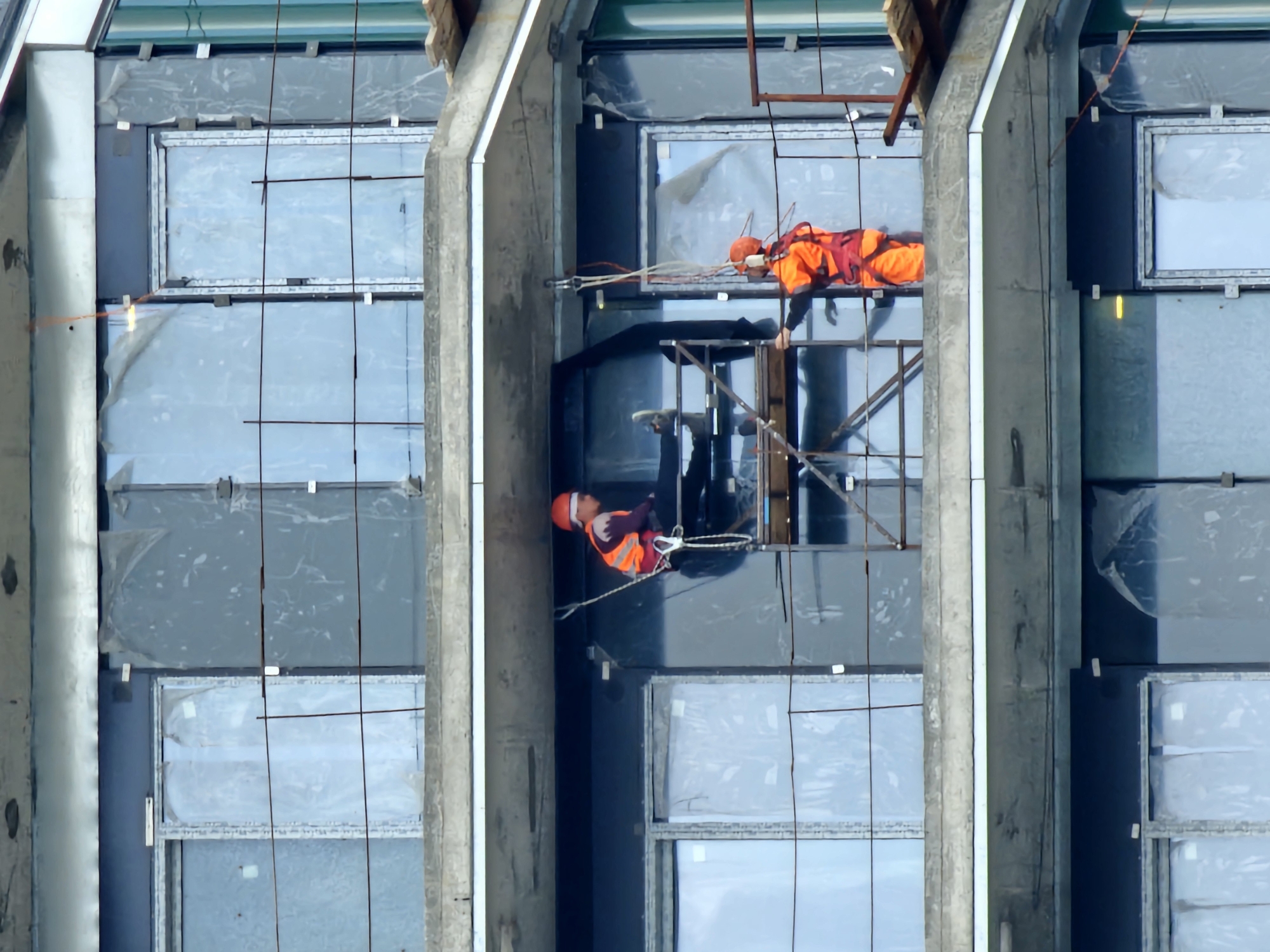A top-down perspective captures two workers suspended on scaffolding between windowed sections of a high-rise building in Moscow, Russia. Both workers are clad in orange safety vests and hard hats. One worker is positioned at the lower level of the scaffolding, while the other is on the upper level. The building's facade is composed of multiple, rectangular glass panels, many covered with a protective film. Concrete pillars vertically divide the windowed sections. The sky is overcast, casting a diffused light on the scene.
