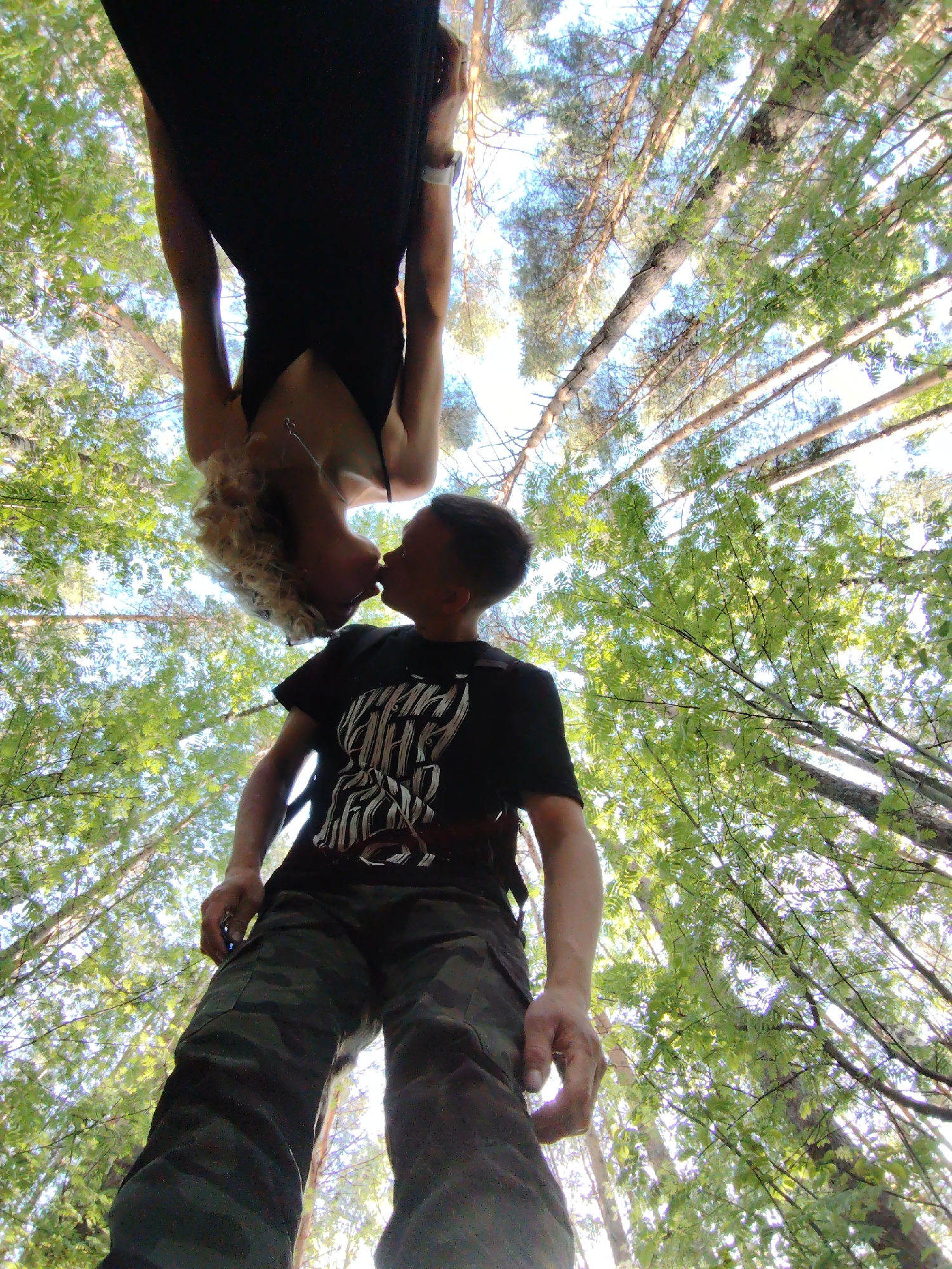 A low-angle, tilted photograph shows a man kissing a woman upside down in a wooded area. The man stands wearing a black t-shirt with white graphics, and camouflage pants. The woman hangs upside down, wearing a black dress. The background is composed of trees with green leaves, and glimpses of a bright sky above. It is not possible to determine the exact location within the city of Chaikovsky, Russia based solely on the image.