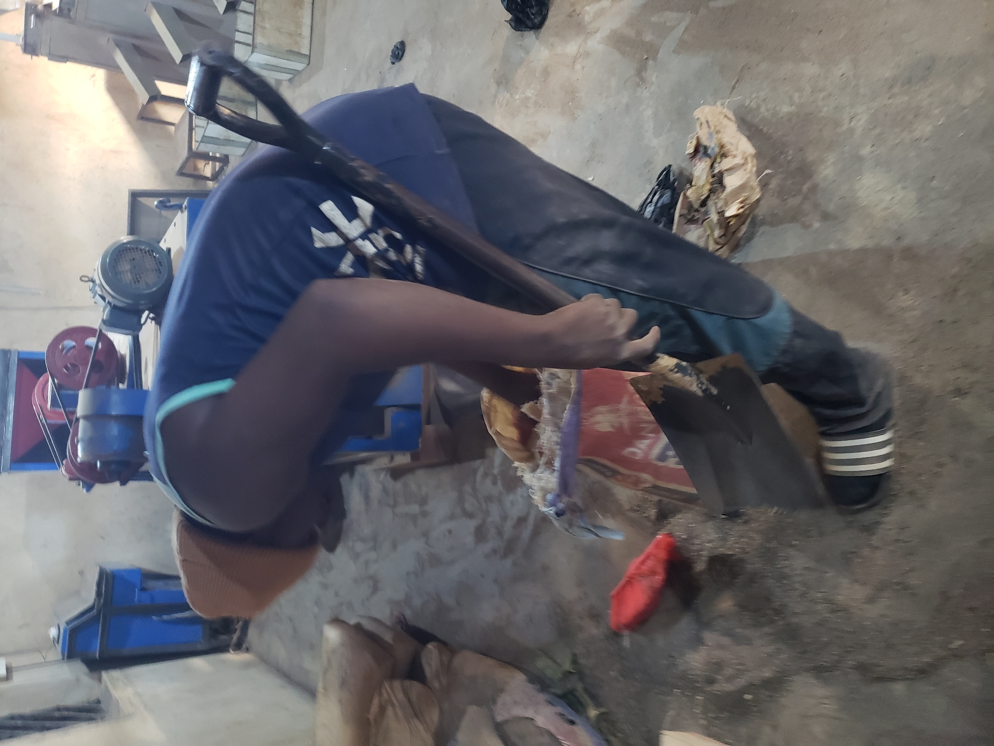 A person wearing a blue shirt with a white logo and dark pants is using a shovel to move material from a red and white bag on a concrete floor. They are also wearing a tan beanie and black and white striped slides. The person is in an indoor industrial setting with various machines and bags of material in the background. The environment appears to be a workshop or factory.