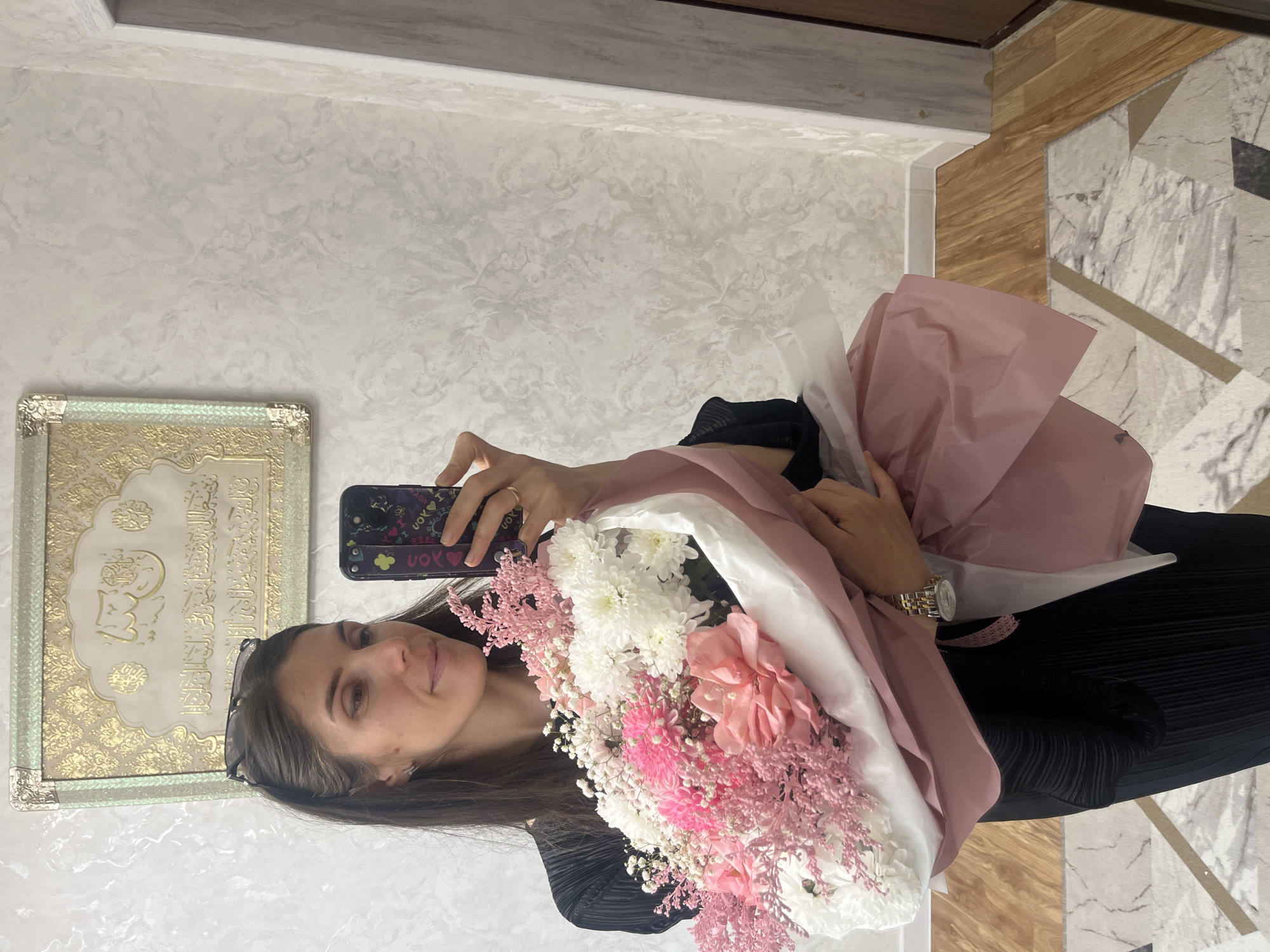 A young woman is captured in a mirror reflection, holding a large bouquet of flowers and taking a selfie. She has long dark hair and a gentle smile, with sunglasses resting on her head. Her left hand holds a smartphone with a dark case featuring "UOY" (YOU reversed) and "LOVE" decals, while a gold-toned wristwatch is visible on her right wrist.
The prominent bouquet displays an assortment of white, pink, and fuchsia flowers, including chrysanthemums and baby's breath, wrapped in soft pink and white paper. She wears a dark, pleated top.
The indoor setting features a light-colored wall with a textured, possibly Venetian plaster, finish. To the left, a decorative framed art piece with ornate gold detailing and Arabic calligraphy hangs on the wall. The floor combines light wooden panels with geometric marble-patterned tiles in shades of white, grey, and black. A grey ceiling trim is visible at the top. The scene is brightly lit, suggesting daytime. The presence of Arabic script hints at the cultural context of the Kaspiysk region.