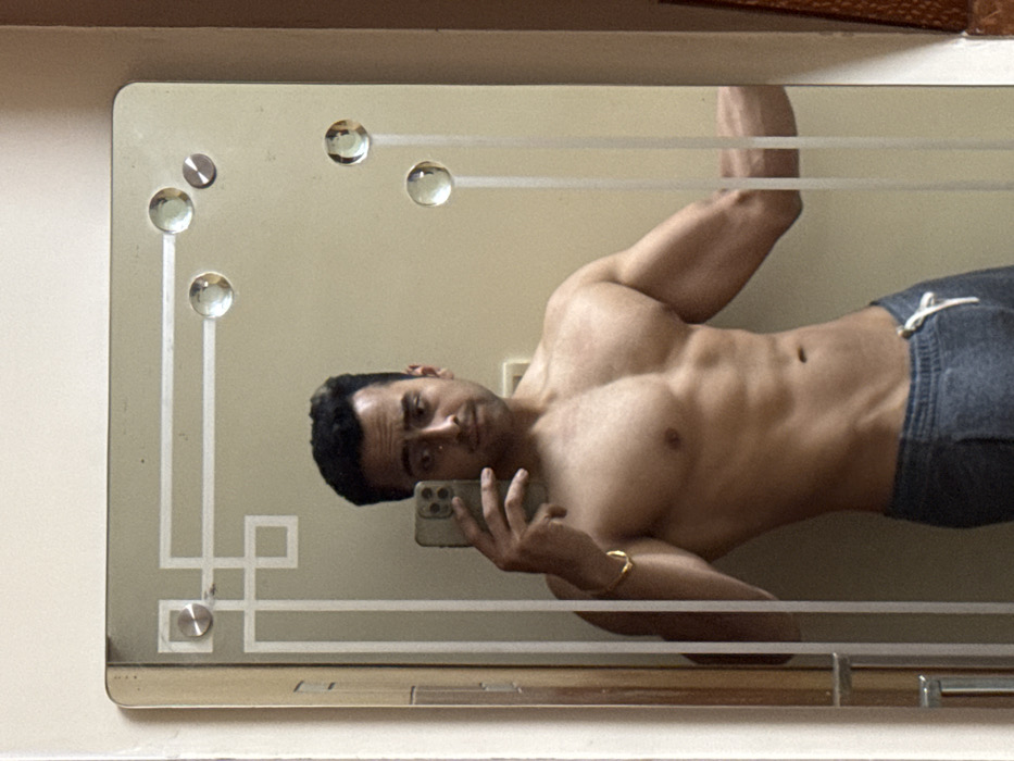 A man with dark hair takes a selfie in a mirror. He is shirtless, revealing a muscular physique. He wears gray sweatpants and a gold bracelet. The mirror has an ornate design with geometric shapes and glass accents. The background wall appears to be a light beige or cream color.