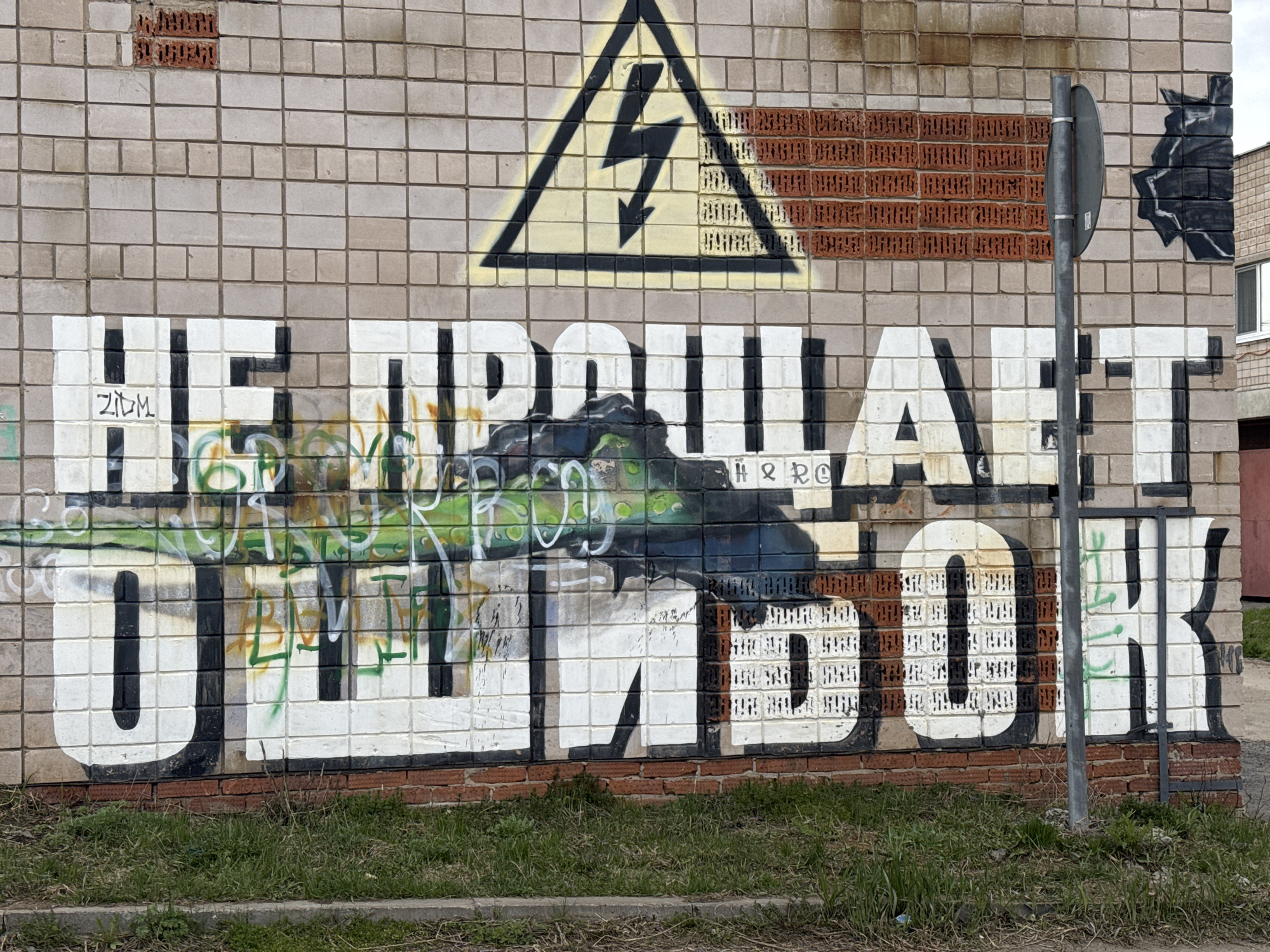 A graffitied brick wall in Zav'yalovo, Russia features an electric shock hazard symbol above a large, stylized Russian phrase "Не прощает ошибок" (roughly translates to "Doesn't forgive mistakes"). The phrase is partially obscured by green and black graffiti which also depicts a tank. A red brick section is intermixed with the gray brick wall, above the phrase. The base of the wall is also lined with red brick, with grass growing along the bottom. A metal pole is visible on the right side of the frame.
