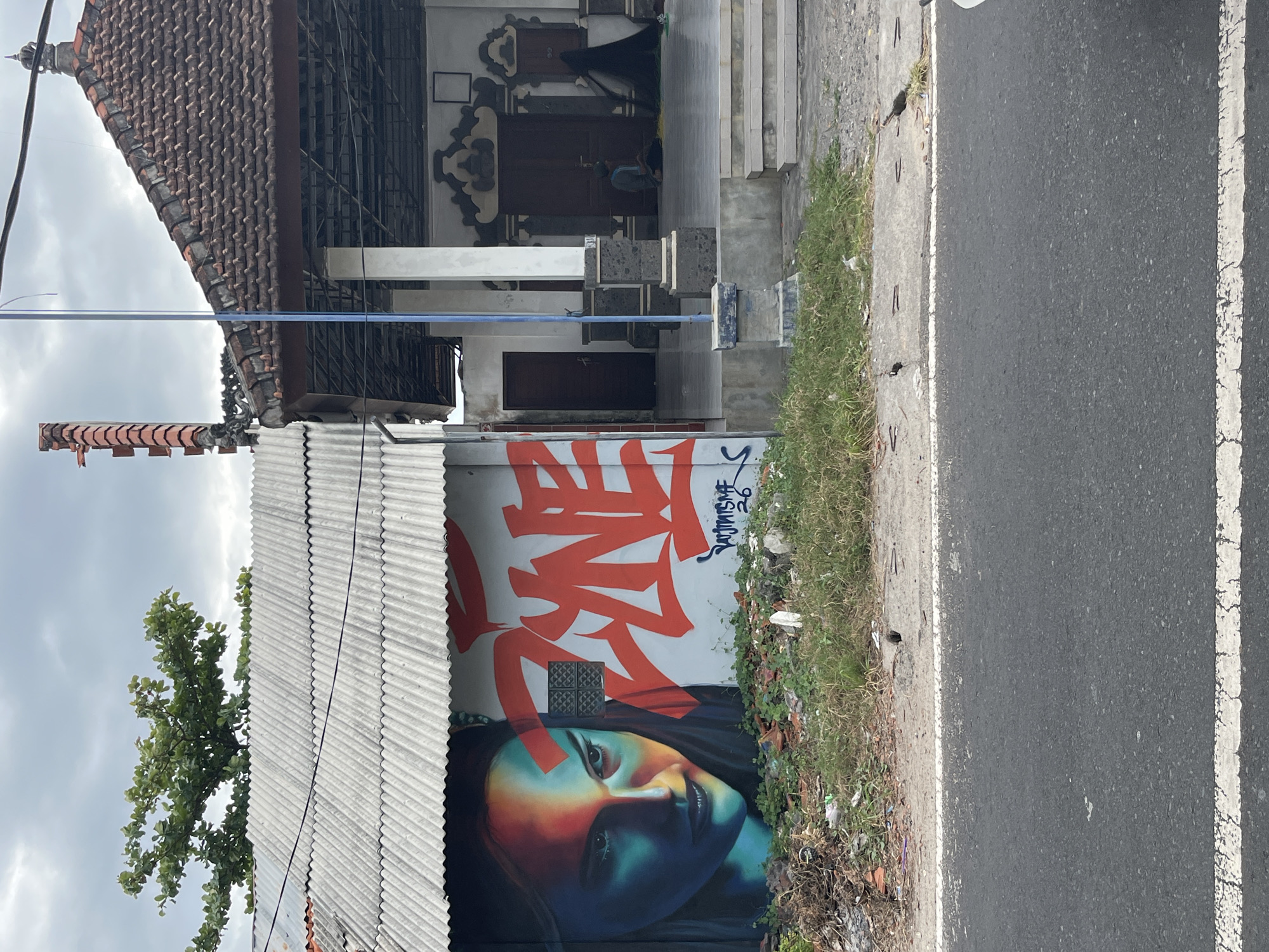 In Kuta Selatan, Indonesia, a weathered, corrugated metal structure stands adjacent to a traditional Indonesian building with a tiled roof. The metal building features a vibrant mural of a woman's face, illuminated by red and blue light, with the word "ZYNE" spray-painted in large red letters next to it. Below the mural, a small patch of grass and debris lines the roadside. The traditional building, with its dark wooden doors and ornate trim, appears to be a place of worship or community gathering. A lone figure is seated on the steps, facing inwards, and a black veil hangs from a pole to the right. A blue utility pole with wires runs vertically between the two structures. The scene is set against a cloudy sky, with a street in the foreground.
