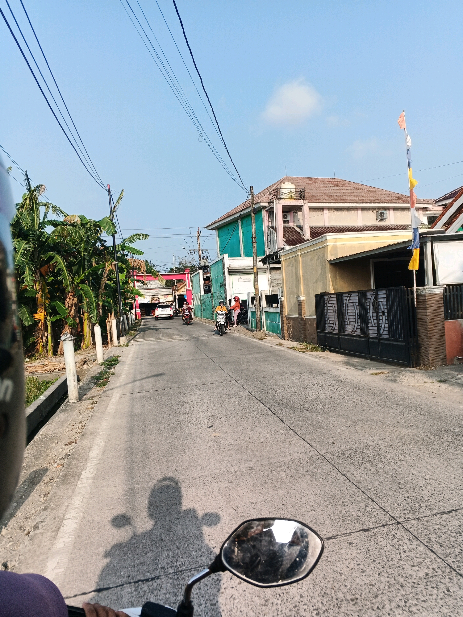 The image depicts a street scene in what appears to be a residential or commercial area with buildings lining both sides of a paved road. The perspective is from someone riding a motorcycle, with a portion of the motorcycle's mirror and handlebar visible in the foreground, along with the rider's shadow on the road. On the left side of the road, there are several banana trees and a concrete drainage ditch. Buildings visible include a multi-story structure with a pale pink facade and an aqua-colored lower section, and a yellow building with a dark metal gate. Several people are on motorcycles traveling down the road, and a white car is also visible further ahead. The sky is clear and blue with a single cloud. Flags are mounted on a pole in front of one of the buildings. The environment suggests a warm, sunny day.