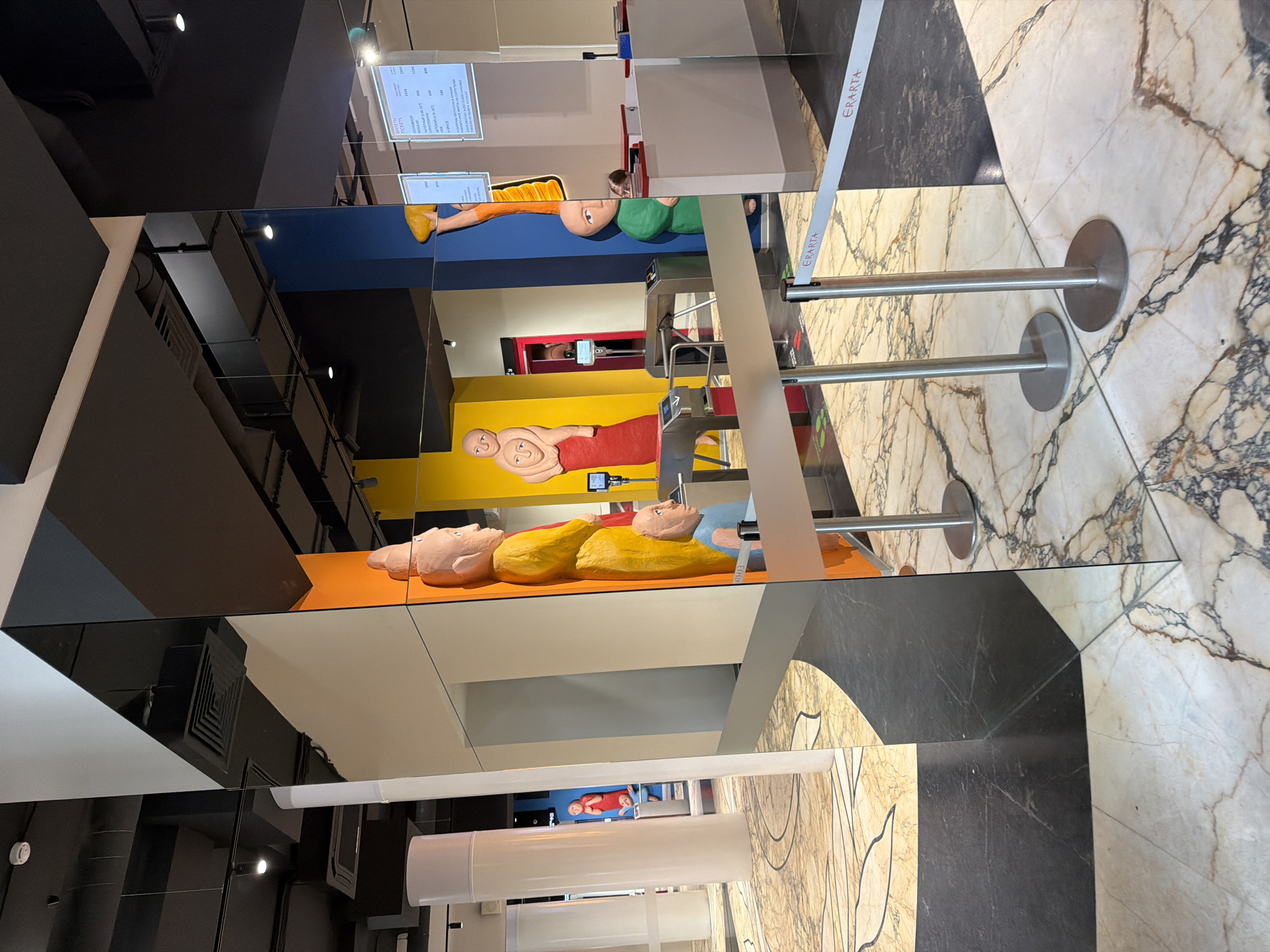 The image captures an interior space at the Erarta Museum of Contemporary Art in Saint Petersburg, Russia. The scene features a display of abstract, reclining human figures painted in bright colors (yellow, orange, red, green, and blue) against corresponding backgrounds. The figures are reflected in a faceted mirror or glass structure, creating a fragmented and multi-perspective view of the artwork. A retractable belt barrier, with the name "ERARTA" printed on it, is visible in the foreground, suggesting crowd control or access restrictions. The floor is composed of marble tiles with intricate patterns, in both black and white or cream shades. A ticket price list is visible on a screen. The overall atmosphere suggests an art exhibition setting with a focus on contemporary sculpture and design.