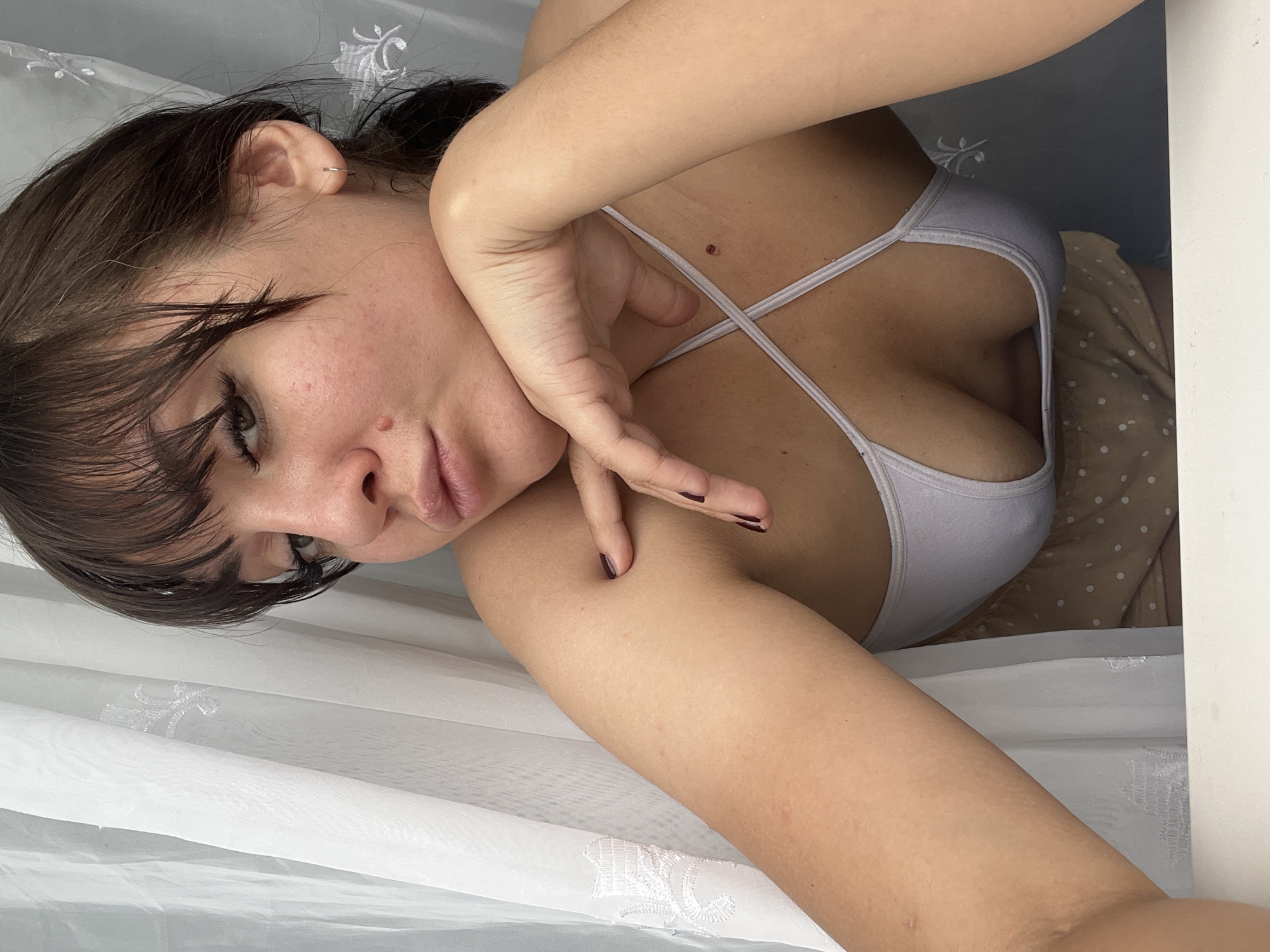 The image shows a person lying on their side, facing the camera. The person has dark hair, parted with bangs that fall across their forehead. Their eyes are looking directly at the viewer with a neutral expression. Their right arm is bent at the elbow, with their hand resting near their face and neck. They are wearing a light-colored, strappy tank top. The background appears to be a bed with white curtains or fabric draped around, and a patterned sheet or duvet behind them. The lighting suggests it is daytime, possibly indoors. There is no visible text in the image.