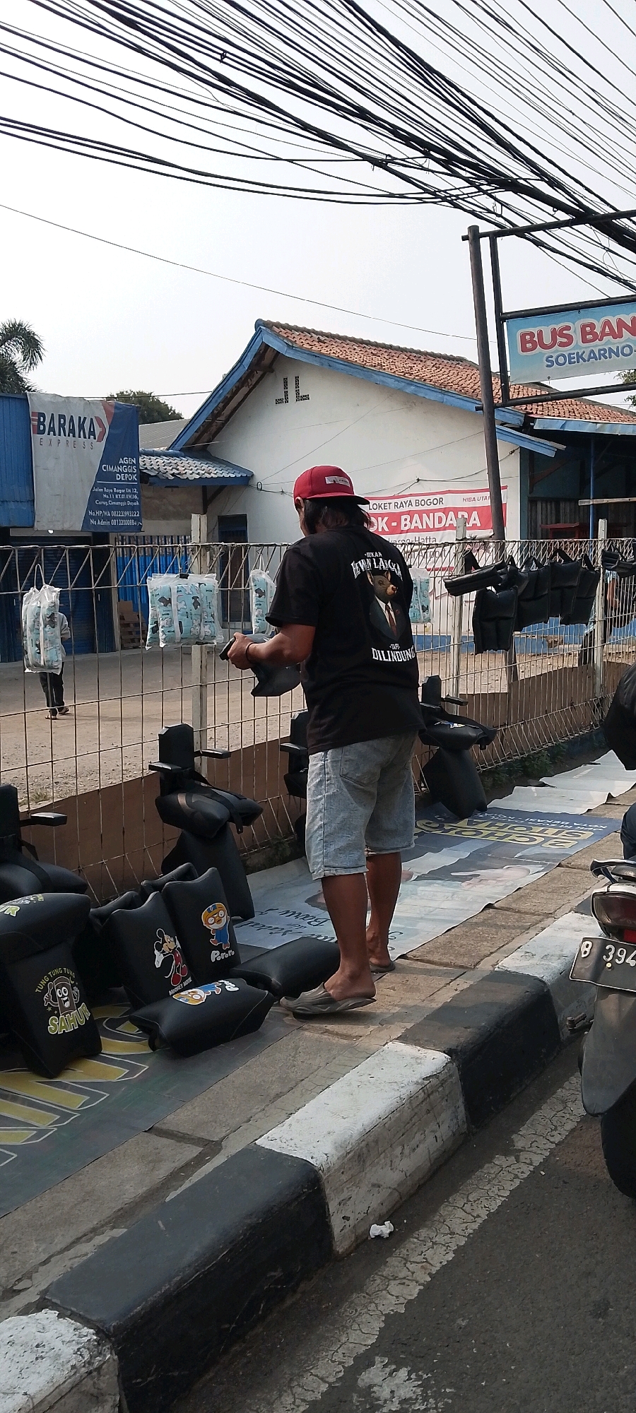A man wearing a red cap and a black t-shirt with an image of a pig in a suit is standing by a fence, holding two black motorcycle seat covers. He is in the process of hanging them on the fence. He is wearing light blue denim shorts and flip-flops. In the foreground, there are several other black motorcycle seat covers, some with cartoon characters printed on them, arranged on the ground and leaning against the fence. Behind the fence, there are buildings with tiled roofs and signs indicating businesses. One sign says "BARAKA EXPRESS AGEN CIMANGGIS DEPOK." Another sign reads "BUS B..." and "SOEKARNO-HATTA". The scene appears to be a street in Depok, Indonesia, possibly a marketplace or a shop selling motorcycle accessories. The environment is outdoors, with overhead wires visible against a bright sky. The curb in the foreground is painted black and white.