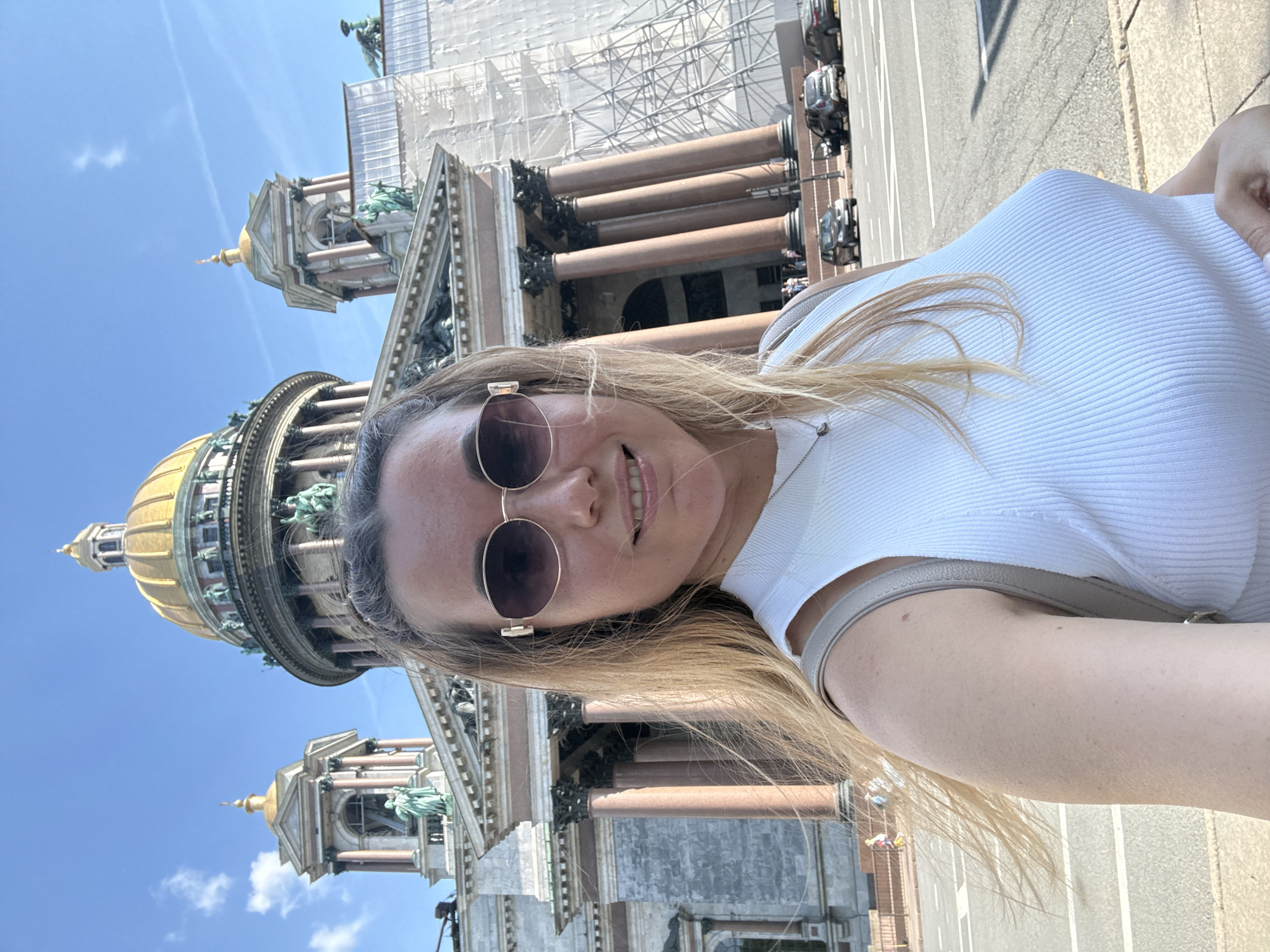 The image, when viewed upright, depicts a smiling woman taking a selfie outdoors on a sunny day, with a grand architectural landmark in the background. The woman, positioned on the right side of the frame, has long blonde hair and is wearing sunglasses. She is dressed in a white, ribbed sleeveless top and has a light-colored strap from a shoulder bag visible on her shoulder. Her expression is joyful, with a wide smile directed at the camera.
Behind the woman, dominating the left side of the frame, is a large, ornate building featuring a distinctive golden dome and a classical colonnade. Numerous statues are visible along the building's facade and around the base of the dome. A significant portion of the building, particularly the upper sections and parts of the columns, is covered in white scaffolding, indicating ongoing construction, renovation, or maintenance. The architectural style, including the golden dome and monumental scale, strongly suggests the building is St. Isaac's Cathedral, likely located in Saint Petersburg, Russia.
The sky is clear and bright blue with a few faint clouds, indicating excellent, sunny weather. The strong light and shadows suggest it is midday or early afternoon. To the right of the building, a paved street with several parked cars is partially visible in the background. There is no visible text in the image.