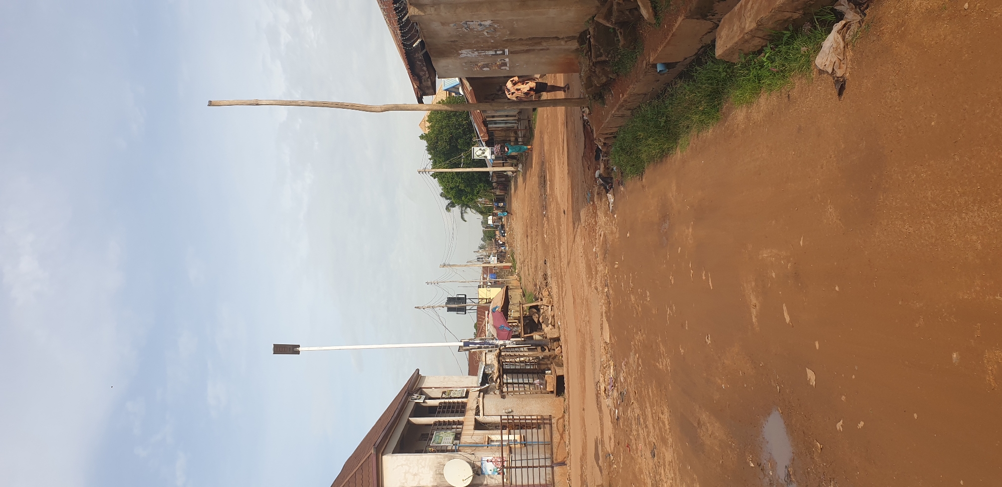 This image depicts an unpaved, reddish-brown dirt road in a semi-urban environment consistent with Sabon Tasha, Nigeria. The time of day appears to be daylight, under a light blue sky with scattered white clouds.
On the left side of the frame, a two-story building with a light-colored facade and a dark brown tiled roof is visible. It features several rectangular windows and a balcony with metal railings on the upper floor, and a white satellite dish is attached to its exterior. In front of this building, a rudimentary wooden stall stands, and a tall, white pole with a dark, rectangular fixture (possibly a solar street light) on top rises prominently. Further down the left side, black water tanks on stands are visible behind other structures.
The road surface is uneven, marked with ruts and depressions, and one distinct puddle of water is present in the foreground on the left. Scattered debris, including pieces of paper and plastic, lies on the road and along its edges.
On the right side of the frame, a single-story building with a rough, unpainted facade and a corrugated metal roof is present. Barbed wire is visible along the top of a wall section, and several faded papers or posters are affixed to the building's wall. A sturdy, unpainted wooden pole stands in the foreground, adjacent to a concrete drain structure filled with green grass.
Several individuals are visible walking along the road away from the viewer. One person on the far right wears a light-colored patterned shirt and dark shorts. Another person in a teal or green garment walks on the left side of the road, carrying an object on their head. In the mid-ground, a black-and-white portrait poster is affixed to a pole or wall. Numerous utility poles with crisscrossing overhead wires line the street, extending into the background, where more distant buildings and sparse trees are visible.