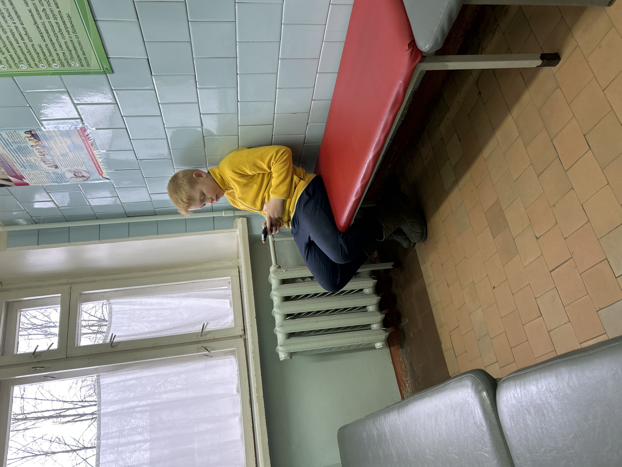 Here's a detailed description of the image:
**Scene:** An indoor waiting area, possibly a clinic or hospital, based on the tile walls and type of seating.
**Location:** Likely in Chaikovsky, Russia, considering the text visible on a poster on the wall is in Russian.
**People:** A single boy, probably in his early teens, is seen sitting on a red bench. He is wearing a yellow long-sleeved shirt and dark pants. He is holding a mobile phone in his hands.
**Objects:**
* Red bench: The boy is sitting on it.
* Window: A window with white curtains is present with bare branches visible outside.
* Radiator: A white radiator is situated below the window, near where the boy sits.
* Seating: Gray seating can be seen.
* Posters: Two posters are fixed on the tile wall with Russian text.
**Actions:** The boy is sitting alone on the bench, looking at his mobile phone.
**Interactions:** The boy is interacting with his phone. There are no other people to interact with.
**Environment:** The room appears to be clean but somewhat dated, with tiled walls and flooring.