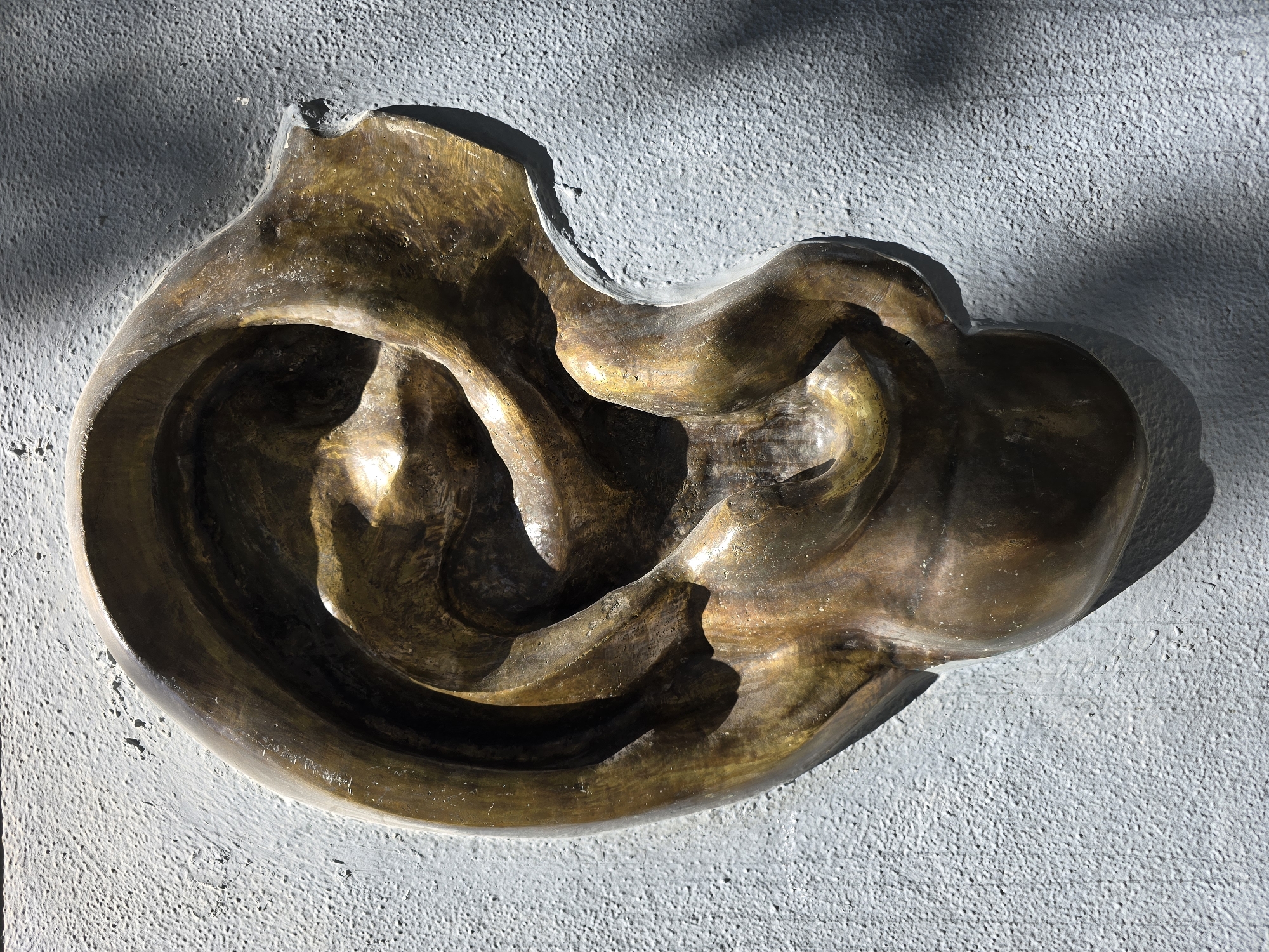 A bronze sculpture of a human ear is mounted on a textured grey wall. The sculpture is detailed, showing the contours and folds of the ear. Sunlight casts shadows on the wall and the sculpture, highlighting its three-dimensional form. The sculpture is located in the courtyard of the Palacio de Justicia in Jaén, Spain.