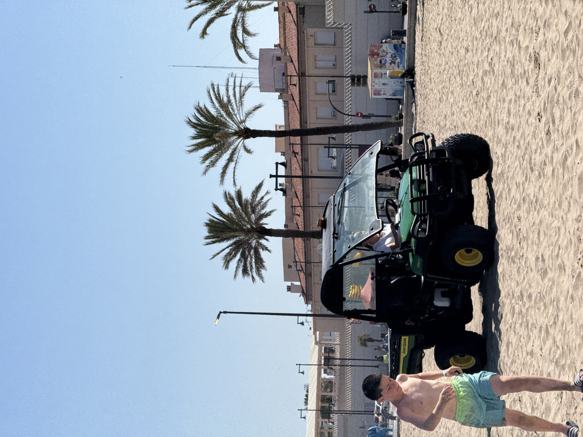 An outdoor, low-angle, full-shot photographic image captures a sunny day on a sandy beach in Valencia, Spain. In the foreground, a young boy, appearing to be in his early teens, walks towards the left of the frame, looking down. He is shirtless and wears teal and light green patterned swim trunks, along with black and white striped flip-flops. To his right and slightly behind him is a green John Deere utility vehicle with a black roll cage and windshield. A person wearing a white shirt is visible driving the vehicle. The vehicle is positioned on the sand, with its front facing the viewer.
In the background, a row of buildings with red tile roofs is visible, situated behind a promenade. Several palm trees are scattered throughout the scene, with clear blue skies overhead. Streetlights with single lamps are present along the promenade. A small, colorful kiosk with graffiti art is situated on the right side of the promenade. The beach is largely devoid of people, except for a few individuals further in the background. The overall scene depicts a typical beach day with recreational activities.
