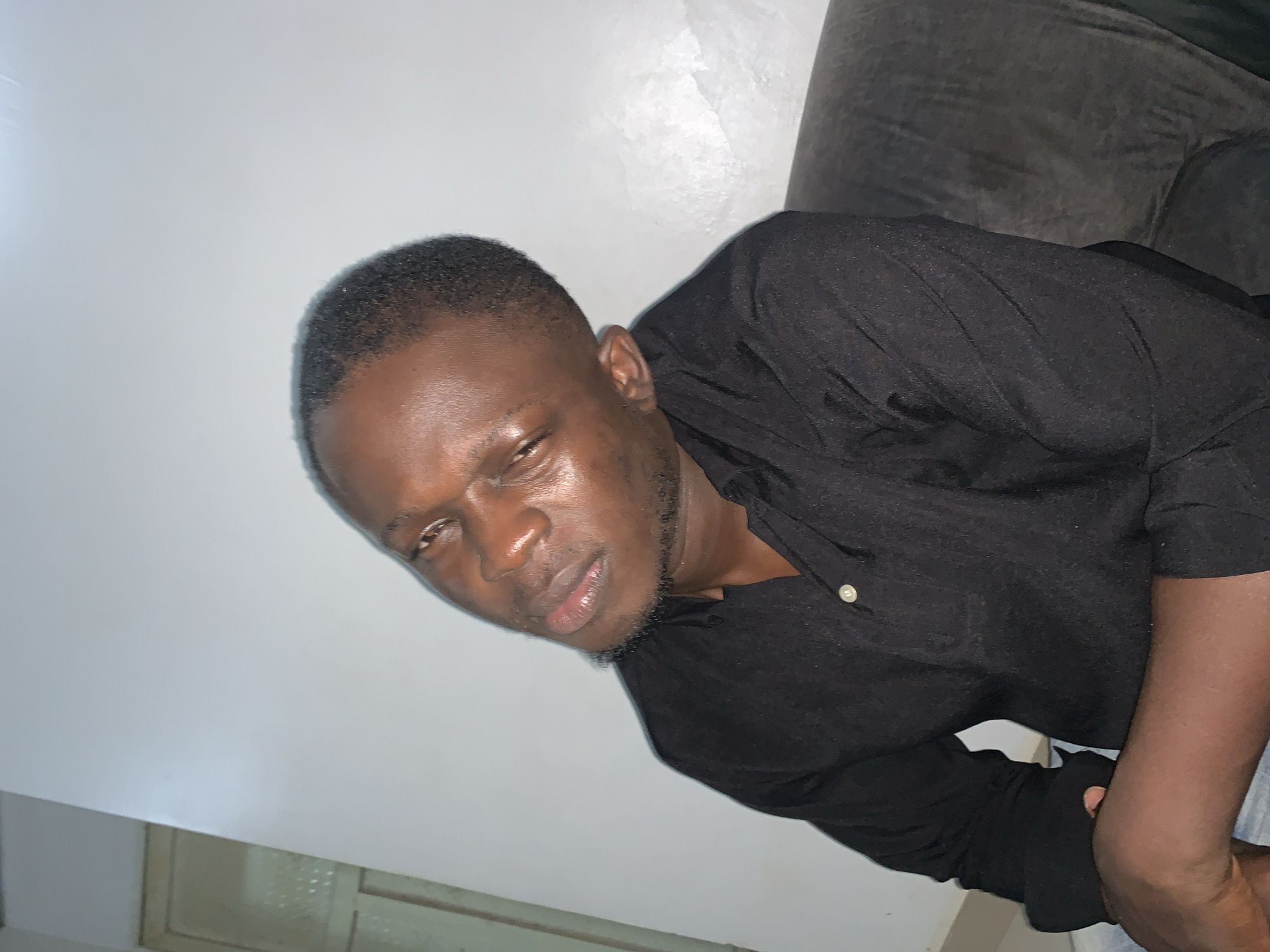 A man with dark skin and short black hair is seated on a dark-colored couch, his head tilted to his left. He is wearing a black collared shirt with a button closure. The background is a plain white wall. There is a doorway visible on the left side of the frame. The lighting appears to be artificial, casting shadows and highlights on the man's face and clothing. The overall scene suggests an indoor setting, likely a residence, in Abuja, Nigeria.