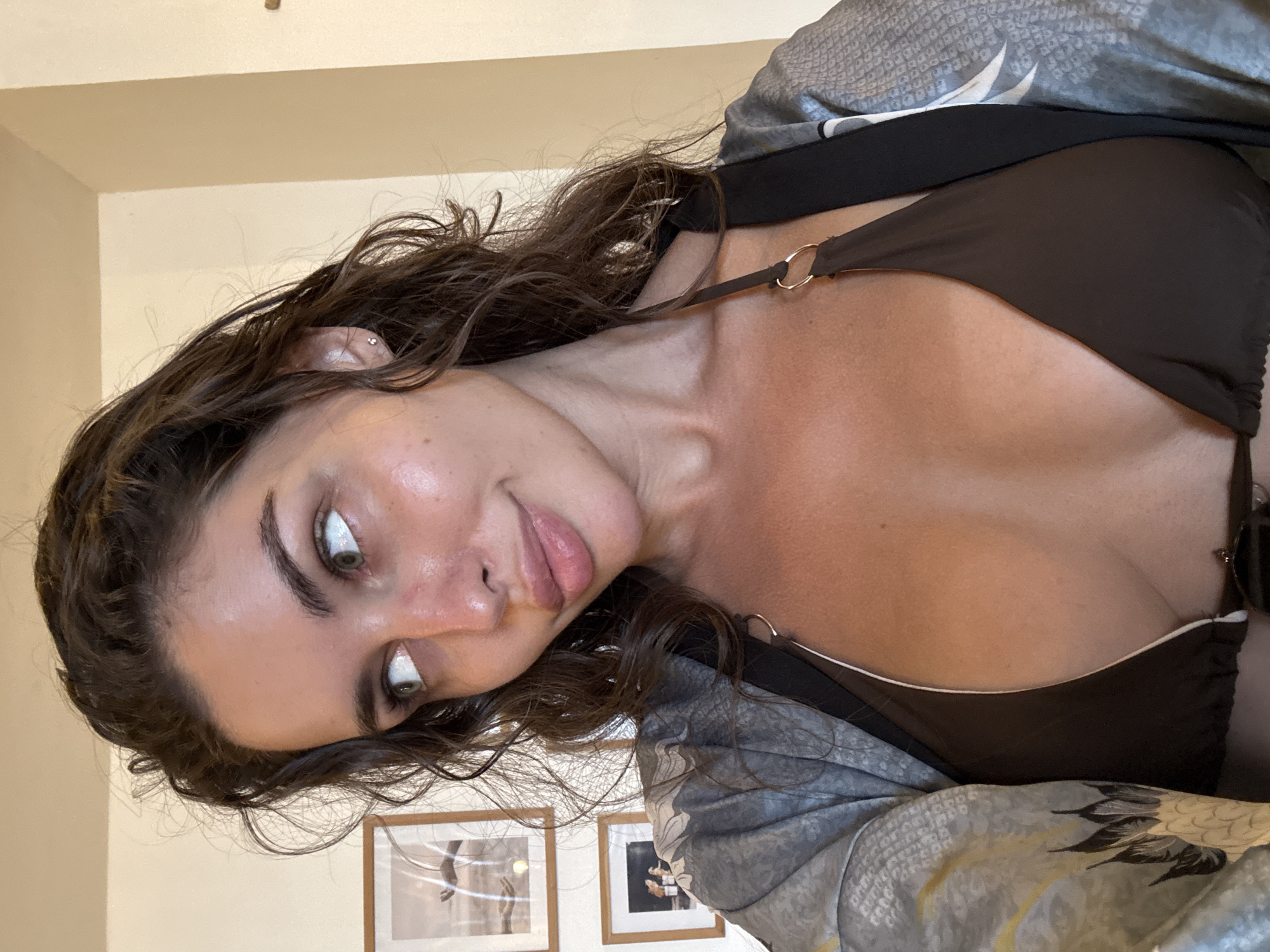 A young woman with light skin, brown hair, and green eyes is photographed from an unconventional angle, tilted towards the left. She is wearing a brown bikini top and a gray patterned robe. Two framed pictures are visible on the wall behind her. The location is unknown, but presumed to be Indonesia, based on the city stated. No specific city details or environmental context are discernible.
