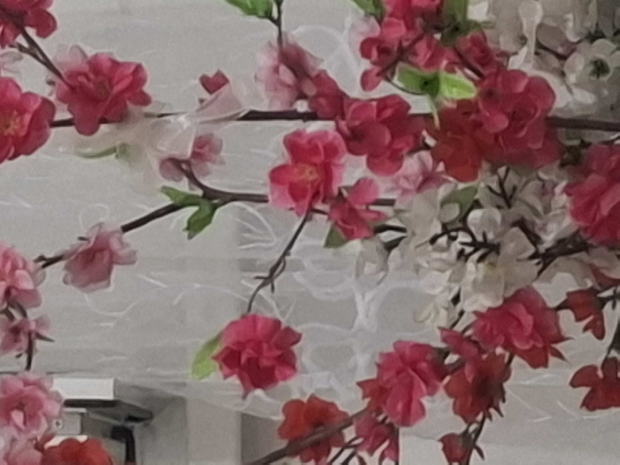 The image displays a close-up view of artificial pink and white flowers, resembling cherry blossoms or plum blossoms, against a white, textured background. The flowers are arranged on dark, thin branches, and some are adorned with delicate white ribbons. The lighting is soft, creating a slightly blurred effect. There are no people or specific city landmarks visible in the image. Therefore, its location in Almaty, Kazakhstan, cannot be determined from the visual information provided. The scene appears to be an indoor decorative arrangement.