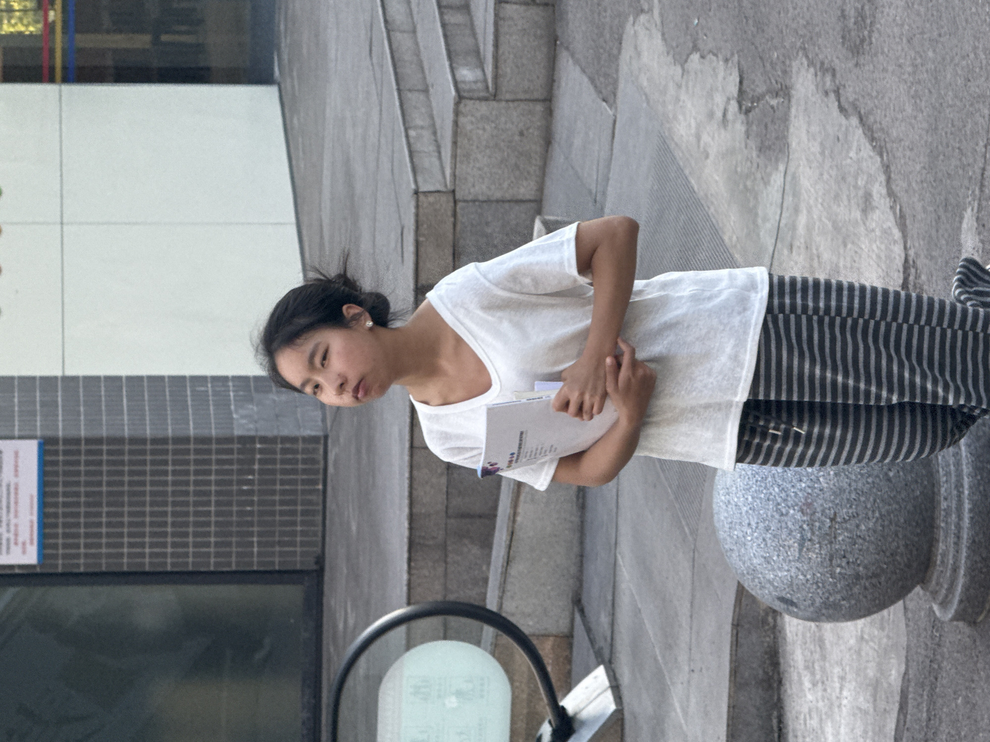 A young woman stands outdoors on a gray stone walkway. She is wearing a white t-shirt and striped pants. She holds a white document in her hands. The location is likely Jiangjin, China, given the presence of Chinese script on a nearby sign. The environment includes steps, a building facade with a poster, and a rounded stone bollard.