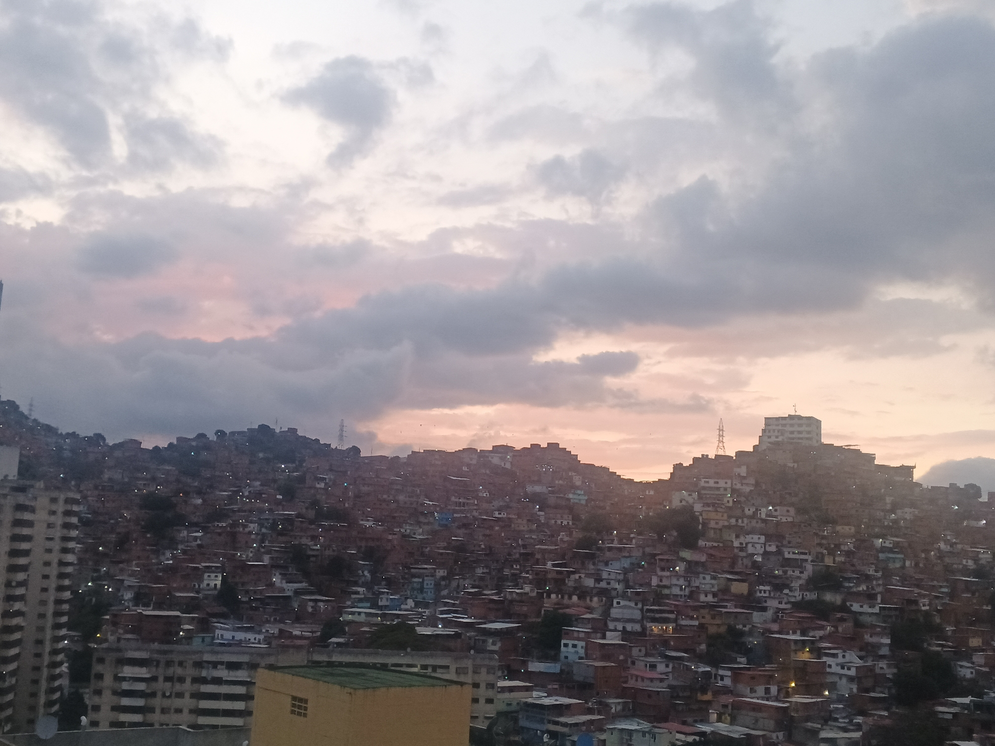 The image presents a panoramic, high-angle view of a sprawling urban hillside settlement under a twilight sky. Thousands of densely packed houses, mostly small and appearing to be informal constructions like shacks or simple brick dwellings, cover the steep contours of multiple hillsides. Many windows within these structures show illuminated lights, indicating habitation as dusk settles.
The sky is a prominent feature, filled with extensive grey clouds. Towards the horizon, particularly in the middle and right sections, a soft pink and orange glow penetrates the clouds, suggesting either a vivid sunset or sunrise.
In the lower left foreground, part of a modern, multi-story apartment building is visible, contrasting with the informal architecture of the hillside. Another lower, yellow-beige building with a green roof is partially seen further to the right in the foreground. A slender transmission tower stands on one of the distant hilltops, and a more substantial, multi-story building crowns another central hill, overlooking the vast expanse of homes. The scene depicts a vibrant yet densely populated urban landscape under a dramatic sky. No visible text is present.