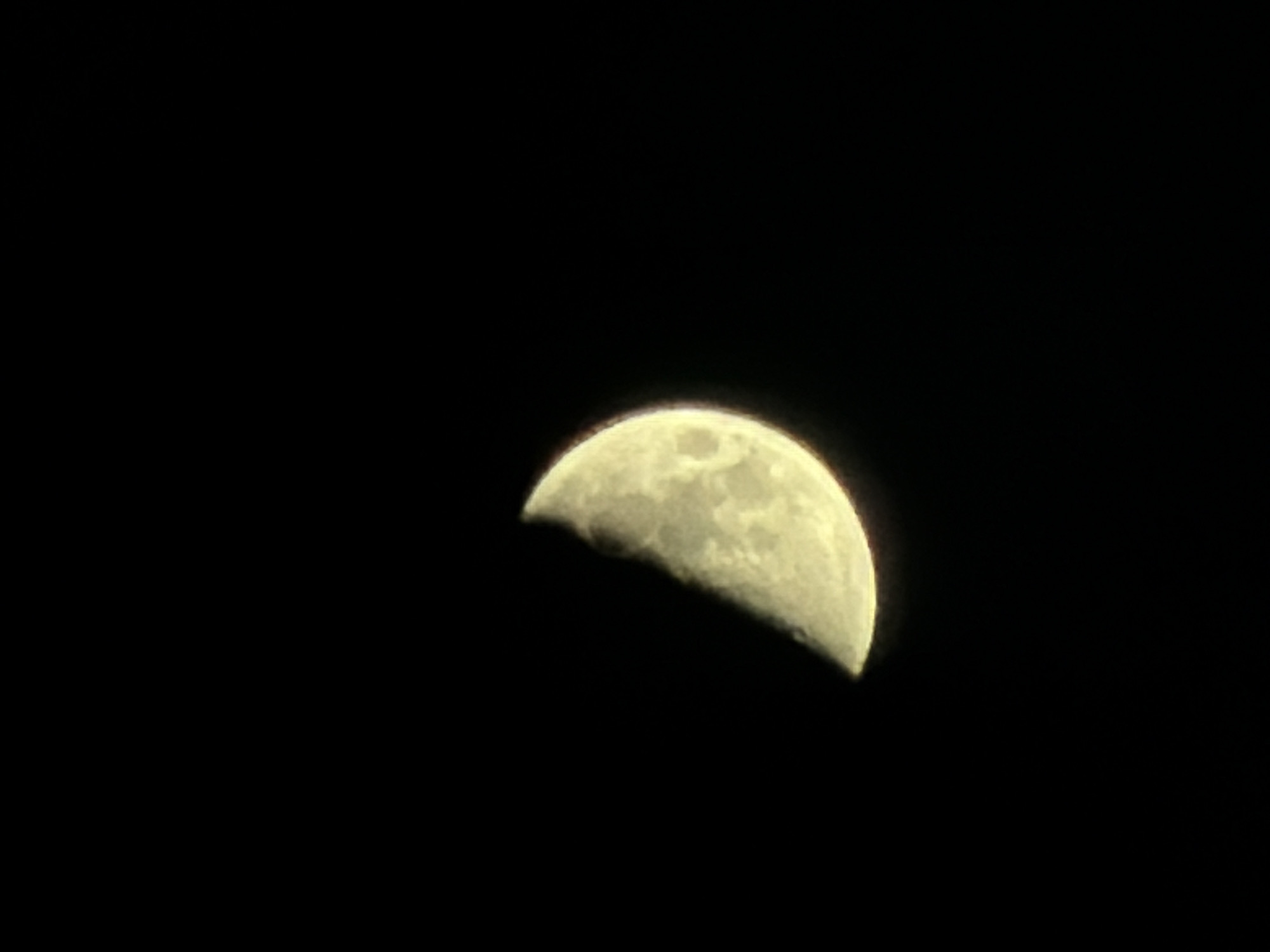 The image presents a detailed view of the Moon against a dark, seemingly cloudless night sky. Approximately three-quarters of the lunar disk is illuminated, indicating a gibbous phase, with its surface appearing in shades of yellowish-white. Observable details on the Moon include various craters and darker regions known as maria, which are typical lunar topographical features.
A prominent, dark, and indistinct silhouette partially obscures the lower-left portion of the Moon. This shadowy element cuts across the bottom of the lunar disc, suggesting a foreground object or natural phenomenon, such as a cloud or distant landscape, positioned between the observer and the Moon. The edges of the illuminated Moon show a slight blur or glow, contrasting sharply with the absolute blackness of the surrounding space.
The scene captures a moment of celestial observation during nighttime. There are no other subjects, objects, or activities depicted, allowing the Moon to be the sole focal point. The dark background confirms the time of day is night, under clear sky conditions allowing for visibility of the Moon. The image does not contain any visible text. Although not evident in the image itself, the provided context indicates the photograph was taken in Burgas, Bulgaria.