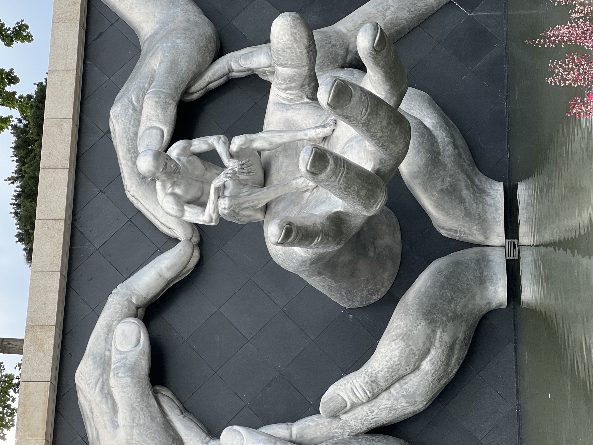 The image shows a sculpture in Krasnodar, Russia titled "Touching Angels". The sculpture consists of giant, silver-colored hands arranged around a central area. Within the hands, there are smaller figures resembling angels in a fetal position. The background is a dark, tiled surface. To the right of the sculpture, there is a water feature with ripples and reflections, possibly a pool or fountain. The hands appear to be made of metal. The sculpture is partially framed by greenery and a light-colored stone structure on the left.