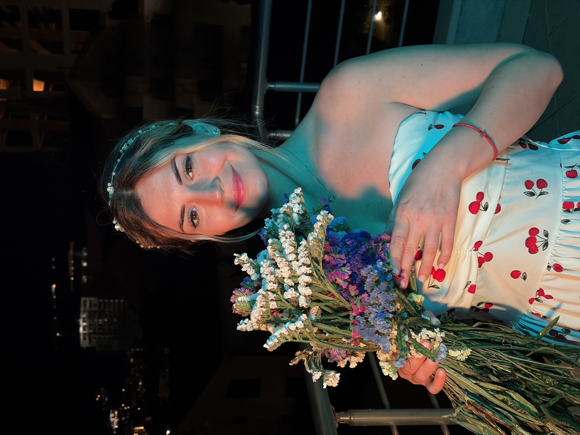 A medium shot captures a young woman holding a bouquet of dried flowers while standing on a balcony at night, with the city of Budva, Montenegro, visible in the background. The woman, with blonde hair styled with a pearl headband, wears a strapless white dress adorned with red cherries and a delicate necklace. She has a gentle smile and is illuminated by warm and cool-toned lights. The dried flowers in her hands consist of various colors, including white, purple, and blue. The balcony railing is visible in the foreground, and beyond it, the cityscape features illuminated buildings and lights. The overall atmosphere suggests a pleasant evening in Budva.