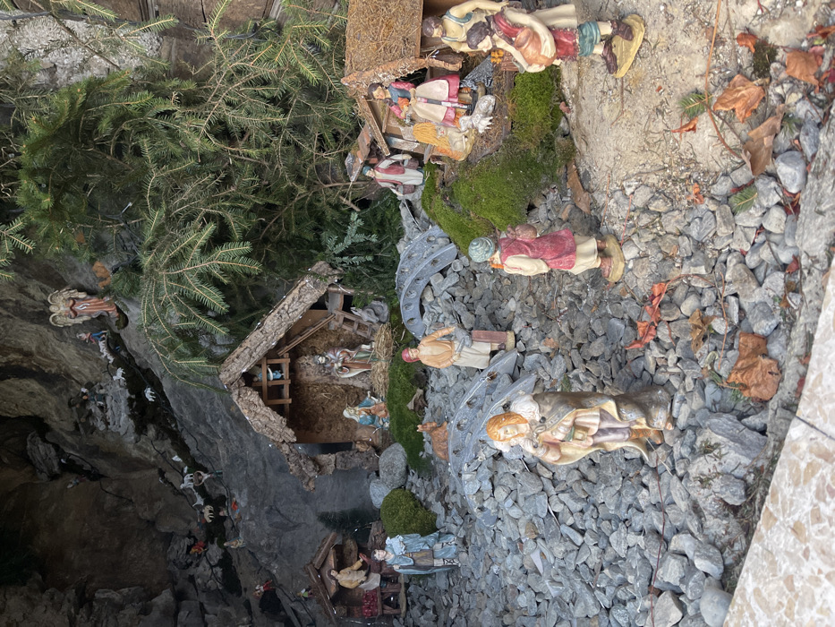This media file captures an elaborate, large-scale outdoor nativity scene, or "presepe," likely displayed in Castione, Italy. The scene is rich in detail and populated with numerous figurines and natural elements, depicting the biblical Christmas story within a rugged, naturalistic landscape.
At the heart of the display is a rustic wooden stable with a thatched roof, where figurines of Mary and Joseph kneel beside the infant Jesus in a manger. Surrounding this central setting, and extending across the rocky ground, are a multitude of other figures. These include angels, some with wings outstretched, shepherds with their flocks of sheep and goats, and the Three Wise Men, all depicted in traditional attire and moving towards or standing near the stable. Additional villagers and various animals, such as donkeys, are also scattered throughout the scene.
The ground is predominantly covered in light grey gravel, suggesting paths and open areas, interspersed with patches of vibrant green moss and scattered dry brown leaves, evoking an autumn or winter setting. Real pine branches and small evergreen trees are strategically placed, creating a natural forest backdrop and enhancing the outdoor feel of the display. To the left, a dark, rough-hewn rocky cliff face forms a significant part of the backdrop, suggesting a grotto or mountainous terrain. Further back and to the left, another smaller, simpler wooden hut with figures inside is visible. The overall impression is that of a meticulously crafted and extensive Christmas display, designed to be viewed outdoors. No visible text is present within the image.