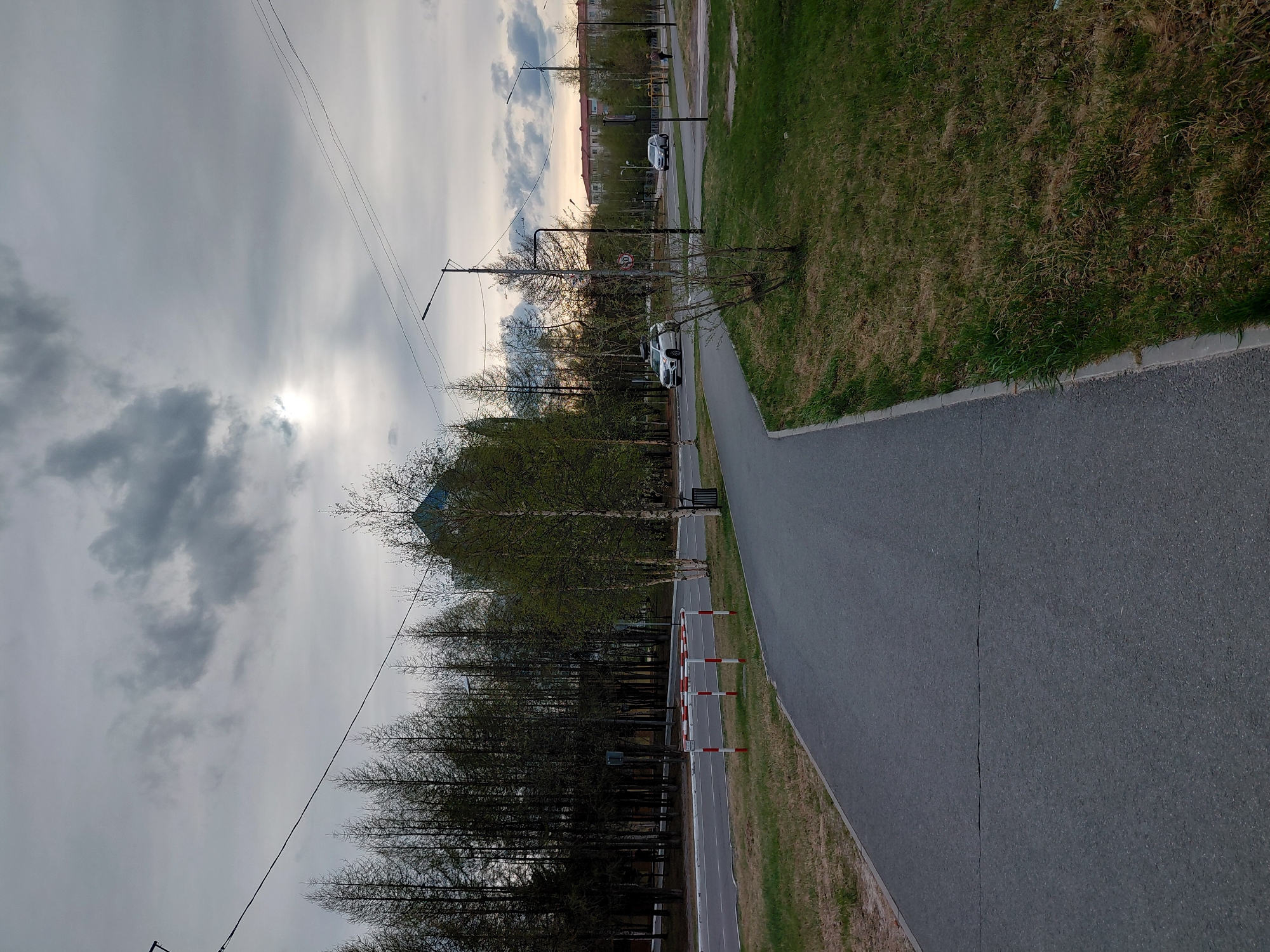 This image presents an outdoor urban or suburban scene, likely in early spring, under an overcast sky with the sun breaking through on the left. The foreground features a wide asphalt path or road, exhibiting a prominent crack running down its center. To the right, a grassy area shows a mix of green growth and drier patches.
On the left side of the main path, a line of deciduous trees stands, some still bare-branched, while others, possibly birches, display the first signs of new green leaves. Beyond these, a narrower, lighter-colored paved path is marked by red and white striped barriers. Several streetlights with horizontal arms rise above the trees, and power lines traverse the sky.
In the mid-ground, a road is visible with two white SUV-style cars; one appears stationary, while the other is further down the road. A circular road sign, likely a "No Stopping" sign (red circle with a blue background and a red diagonal line), is mounted on a pole near the trees. In the distant background, multi-story residential buildings are visible, suggesting an urban environment in Nadym, Russia. The sky is mostly covered with grey clouds, creating diffused lighting, though a bright patch reveals the sun's position. No people are present.