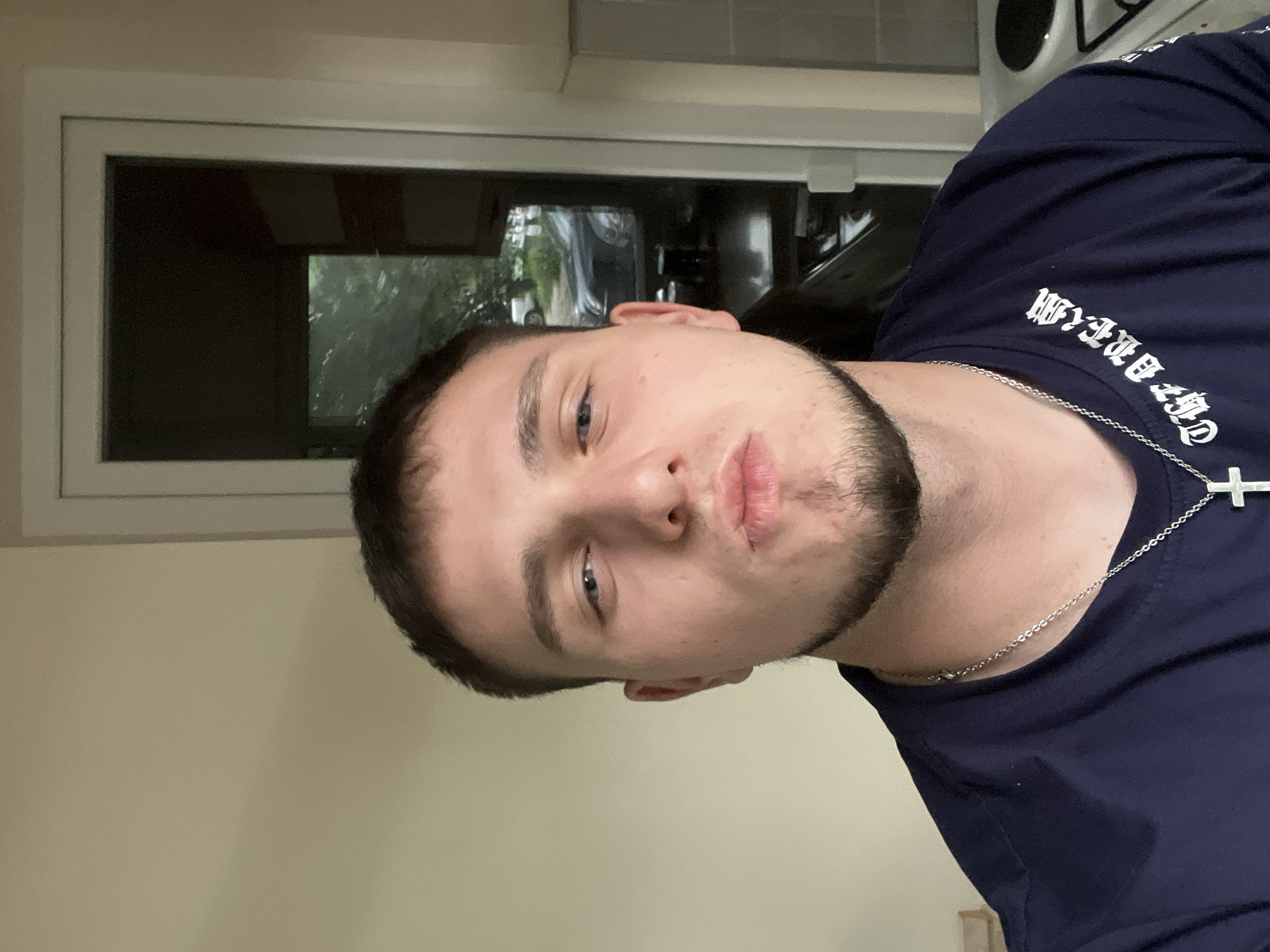 A selfie image shows a young male with short dark hair, fair skin, and a short beard. He wears a dark blue t-shirt with light-colored gothic script and a silver chain necklace with a cross pendant. He appears to be indoors, near a window. Visible through the window are trees, a car, and possibly other buildings. It is not possible to determine if the location is in Plovdiv, Bulgaria based on the information presented in the image.