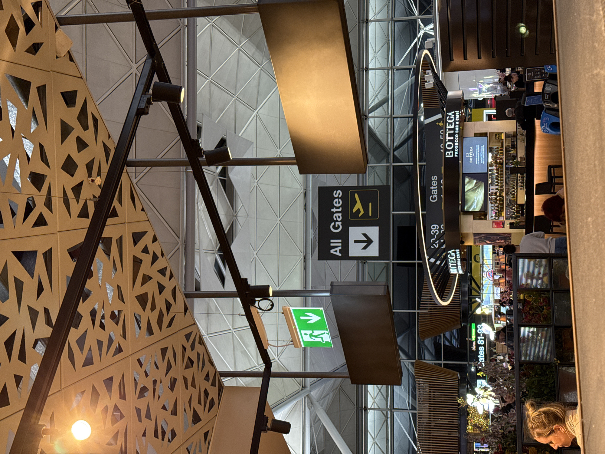 This image captures an interior view, likely within an airport terminal in Uttlesford, United Kingdom. The visual focus is on architectural elements like patterned ceiling panels and a metal support structure with attached spotlights. Overhead signage indicates directions towards "All Gates" and specific gate numbers (81-93, 20-39). A bar/cafe named "Bottega Prosecco Bar & Caffe" is visible, displaying merchandise and serving customers. People are present, including staff behind the bar and patrons, though their actions are not clearly defined. The environment is modern with a high ceiling and large glass windows.