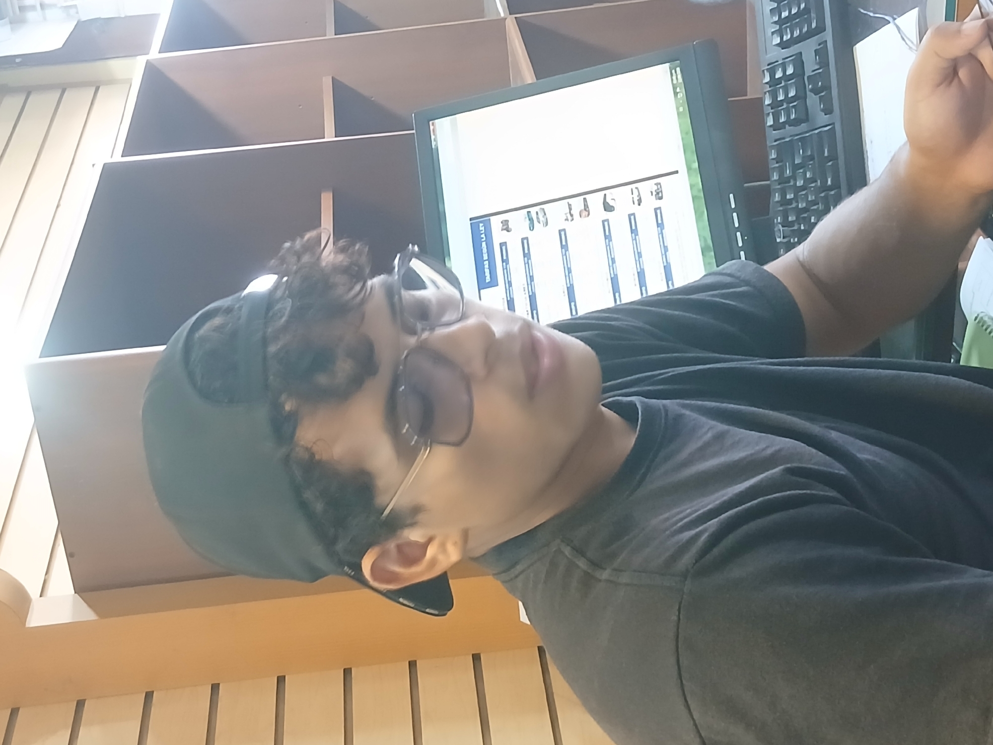 A young male wearing a black baseball cap and a dark t-shirt is seated in front of a computer monitor. The monitor displays a list with images, possibly of vehicles, and text in Spanish that reads "TARIFAS SEGUN LA LEY". A black keyboard is positioned in front of the monitor. The background shows shelving units. The lighting suggests an indoor setting. The image appears to be taken in Juan Griego, Venezuela.