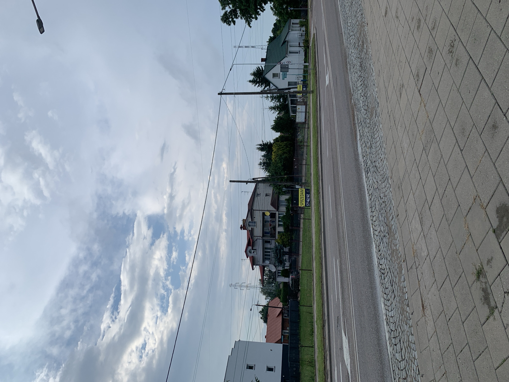 The image shows a street scene in Białystok, Poland. A two-lane asphalt road runs horizontally across the foreground. The sidewalk next to the road is paved with grey concrete tiles. A cobblestone border separates the sidewalk from a strip of grass. Behind the grass, a fence encloses several houses and trees. One house, on the right side of the image, is white with a green roof. Another house, further back and to the left, is a multi-story building with a red roof. A "SPRZEDAM" (For Sale) sign is visible in front of the fence. Overhead, power lines stretch across the frame, and a street lamp is visible in the upper right corner. The sky is cloudy, with patches of blue visible. A large power pylon can be seen in the distance.