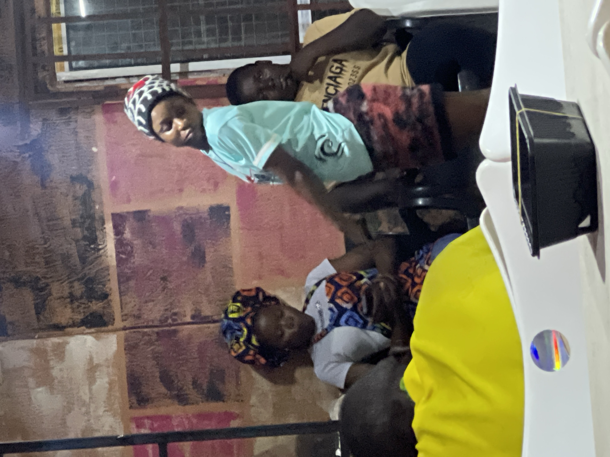 The image shows a group of people seated in an indoor space in Obosi, Nigeria. A woman wearing a patterned headscarf, white top, and patterned skirt is on the left side of the frame. Another woman in a black and white hat and a light blue shirt is seated to the right, while a man in a beige shirt is behind her. A person wearing a yellow top is to the bottom right. There are white plastic chairs in the scene, along with a black object attached to one of the chairs. The background features a textured wall.