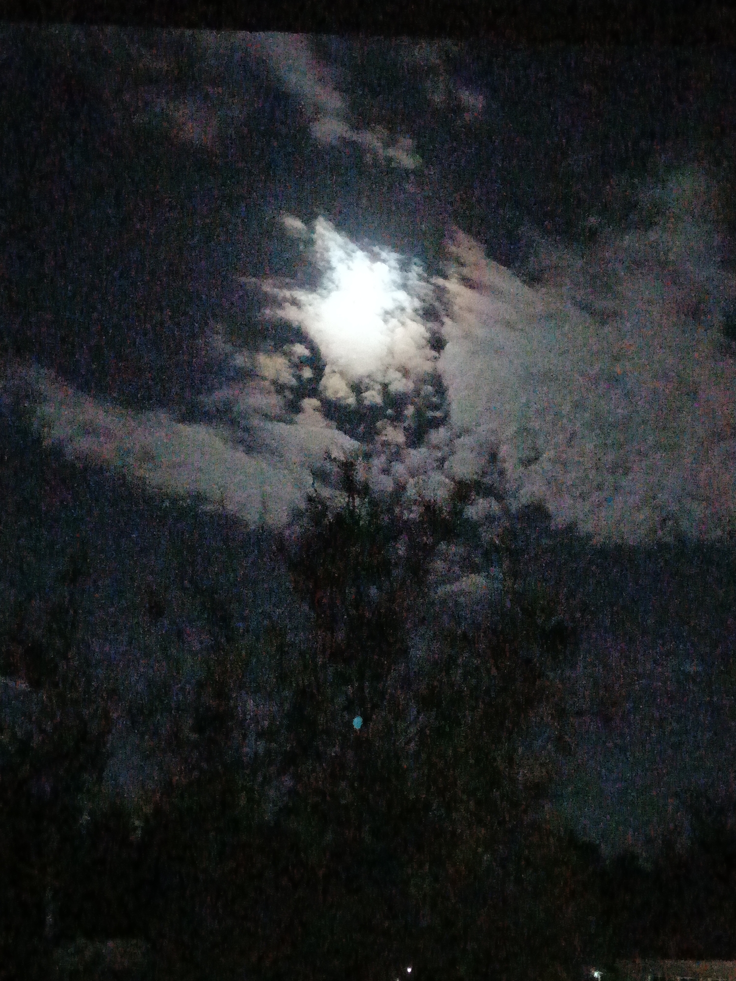 The image captures a dark night sky dominated by a brightly shining moon, partially obscured by clouds. The moon, a brilliant white light source, is positioned in the upper-middle of the frame, emitting a strong glow that illuminates the surrounding clouds. These clouds exhibit varying shades of white and gray, creating a textured canopy across the sky.
Below the cloud-covered sky, the dark silhouettes of trees or dense foliage are visible, appearing as indistinct masses that stretch across the lower and middle sections of the image. Their dark forms contrast with the moonlit sky, enhancing the nocturnal atmosphere. A small, distinct teal-colored dot is visible in the lower-middle area, possibly a lens flare or a sensor artifact.
The photograph has a grainy texture, typical of low-light conditions, which contributes to the moody and mysterious ambiance of the scene. The time of day is clearly night, with the moon being the primary light source. The setting, located in Парэчча, Belarus, appears to be a natural outdoor environment, likely rural or suburban, given the presence of trees and the absence of noticeable artificial lights. There is no visible text in the image.