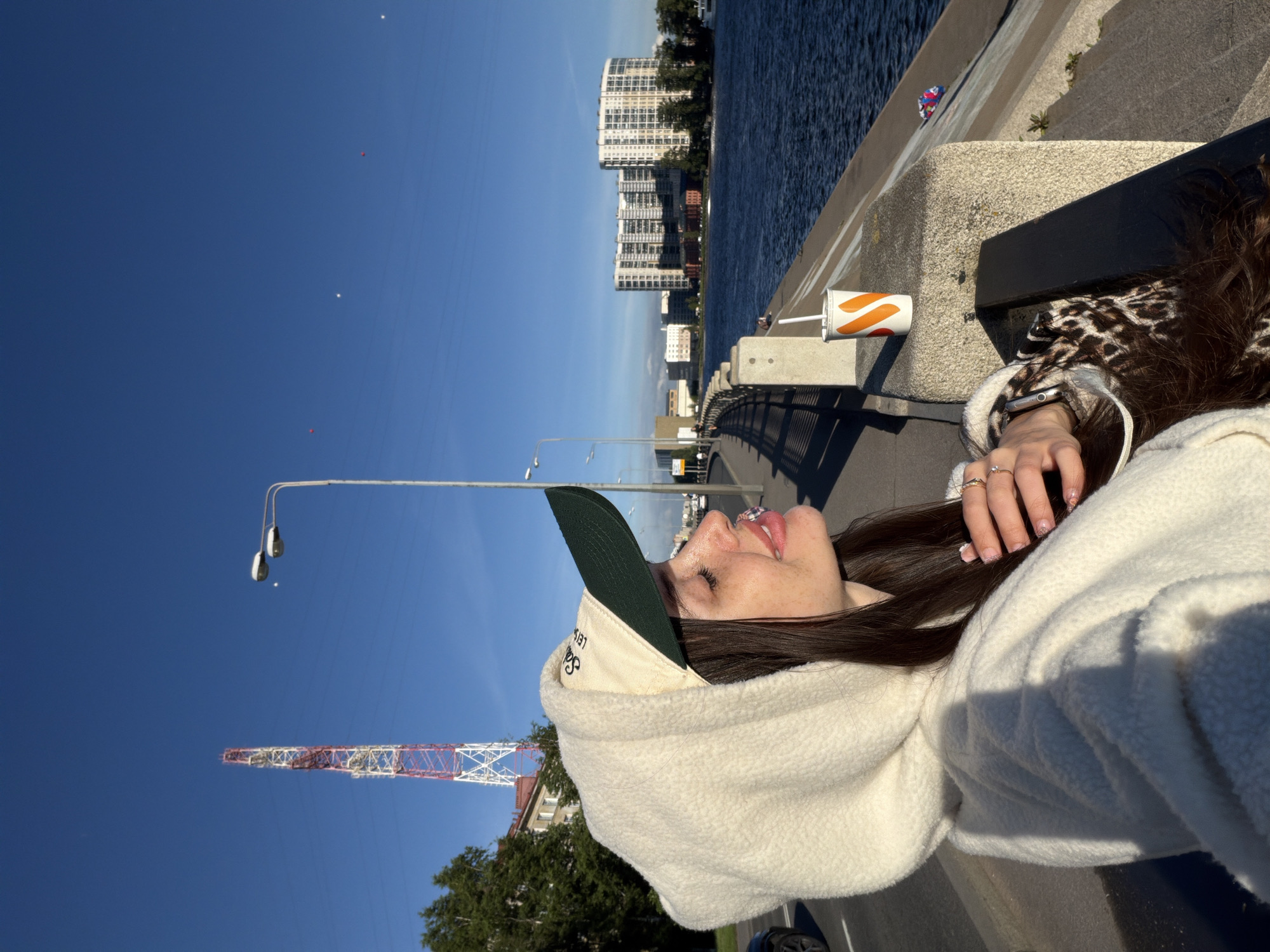 In Shushary, Russia, a young woman with long, dark hair and freckles is captured in a close-up profile shot. She is wearing a cream-colored hooded fleece jacket and a baseball cap with a dark green brim and a light-colored crown. Her eyes are closed, and her face is tilted upwards, seemingly enjoying the sunlight. A disposable cup with orange and white markings, likely containing a beverage, sits on a concrete ledge to her right.
The background reveals a city scene with a wide body of water, possibly a canal or river, on the right. Across the water, several modern, multi-story buildings are visible, reflecting in the calm blue water. A prominent red and white lattice tower, likely a radio or telecommunications tower, stands in the middle distance on the left. A paved walkway with streetlights lines the waterfront. The sky is a clear, vibrant blue, with a few small, distant objects that appear to be balloons. The overall lighting suggests it is a sunny day, with shadows cast on the walkway and the woman.