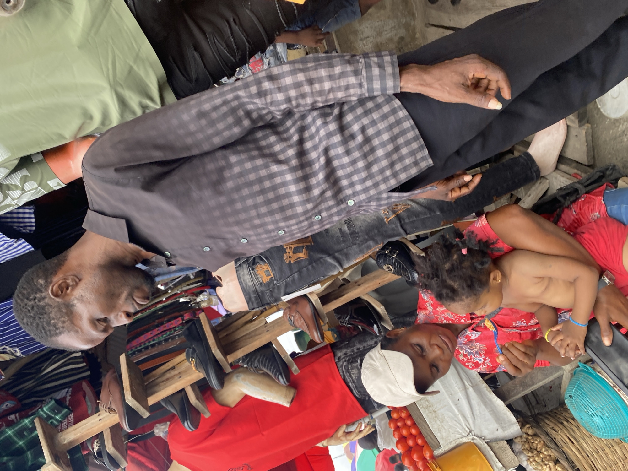 In a busy marketplace in Ado, Nigeria, a man wearing a checkered long-sleeved shirt and black trousers sits with one leg crossed over the other. Near his legs is a woman wearing a white cap and red top is holding a shirtless child with curly hair and colorful bracelets. The child appears to be looking at or reaching towards something in the woman's hand. In the foreground, there's a rack displaying belts and shoes. A variety of items like tomatoes, a blue basket, and peanuts are also visible, suggesting a market scene. The environment appears crowded with various goods and people.