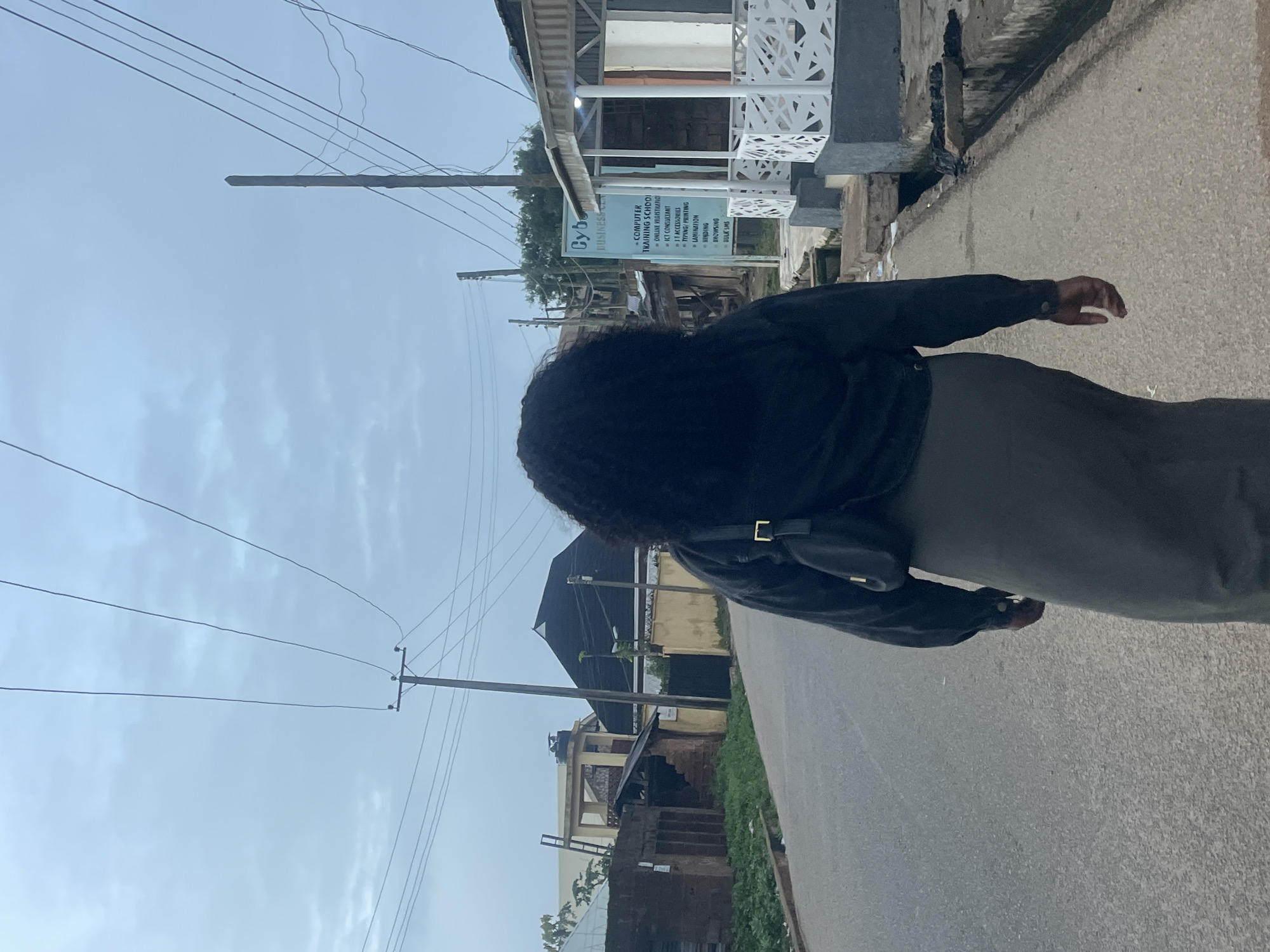 The image depicts a rear view of a woman walking down a paved road in Jos, Nigeria. She has dark, curly hair and is wearing a dark jacket over a long, olive-green skirt. A small black bag is slung across her chest. The road appears to be a typical street in a residential or commercial area, with buildings lining one side. On the right side of the road, a sign for "Cyber Business Centre" is visible, listing services such as "Computer Training School," "Online Registration," "ICT Consultant," "IT Accessories," "Typing/Printing," "Lamination," "Binding," "Browsing," and "Bulk SMS." Utility poles with numerous electrical wires crisscross the overcast sky. The environment suggests a somewhat developed but possibly aging urban setting. The other side of the road features various structures, including brick buildings, a pitched roof structure, and greenery. A drainage ditch runs alongside the road.
