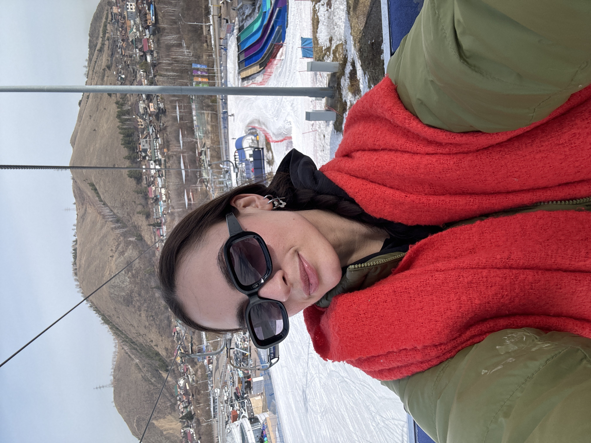 A medium shot depicts a person with fair skin, wearing sunglasses, a red scarf, and a green jacket. The background shows a ski resort landscape. In the distance are buildings and a bare mountain. There are ski lifts and snow-covered slopes. The photograph appears to be taken from a ski lift, looking down at the resort. Based on the distinct mountain profile and colorful waterslides, the location is likely the Bobrovy Log ski resort in Krasnoyarsk, Russia.