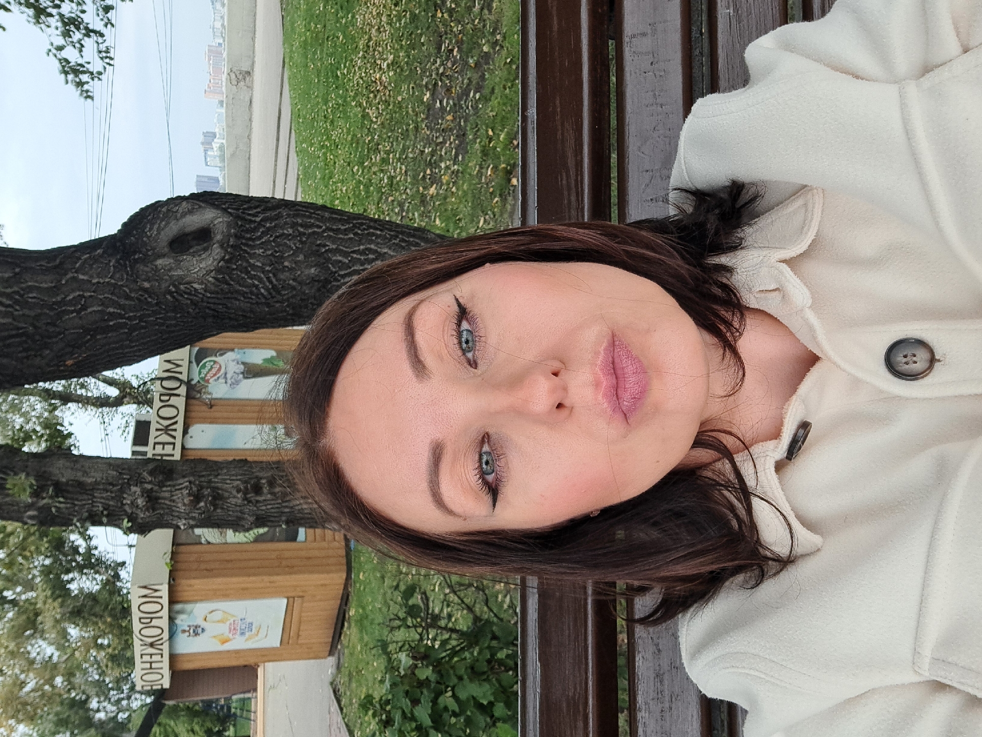 A woman with dark brown hair and blue eyes takes a selfie while sitting on a wooden park bench. She is wearing a cream-colored jacket. Her lips are pursed. Behind her, a wooden kiosk with Cyrillic lettering is visible, and beyond that, a cityscape with buildings and a body of water. The setting appears to be a park in Irkutsk, Russia, indicated by the Cyrillic signage on the kiosk. The ground is covered with fallen leaves, suggesting an autumn setting.