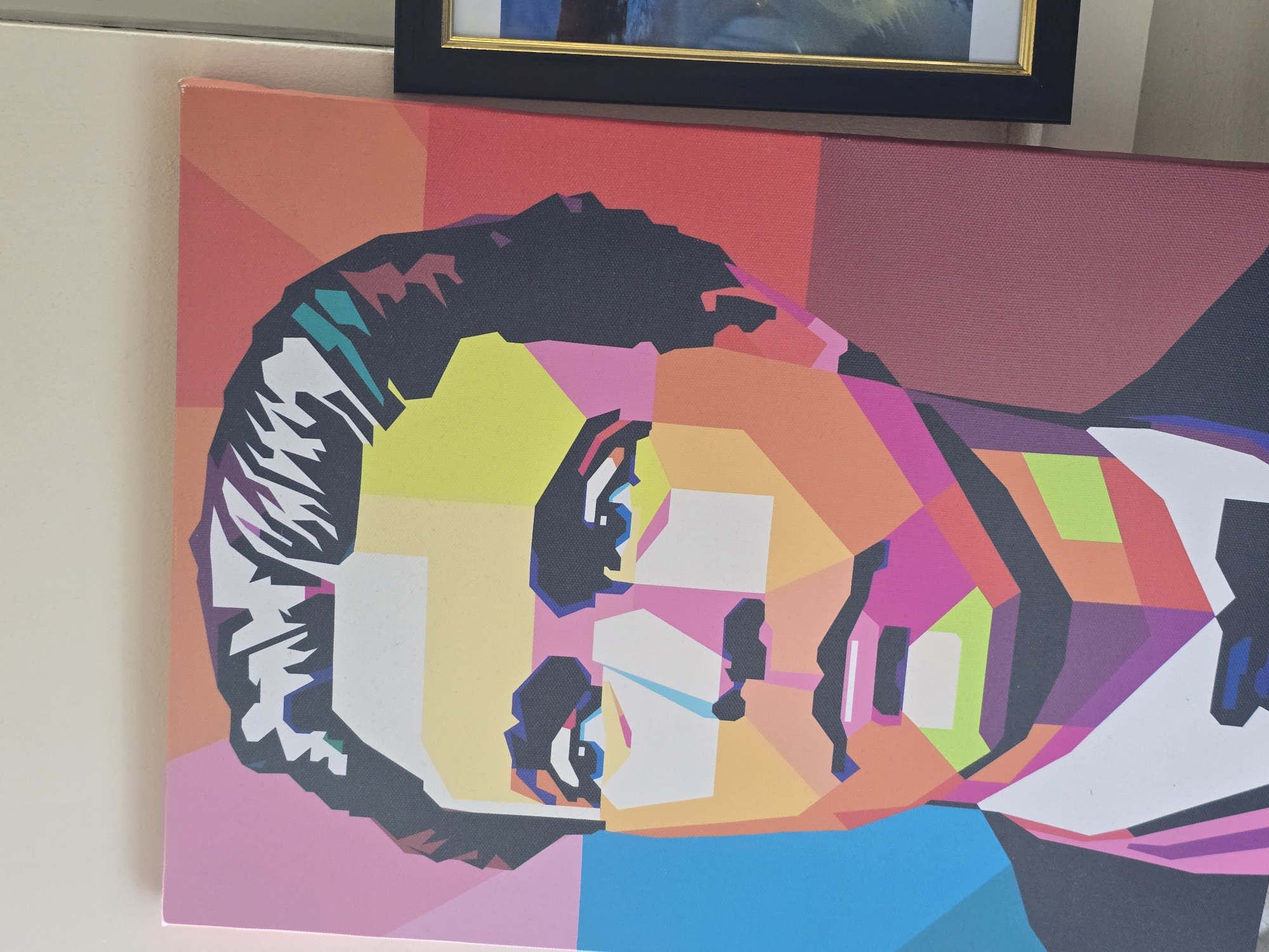 The image displays a stylized pop art portrait of a man, rendered in geometric shapes and vibrant colors. The subject is a close-up shot, focusing on the upper torso and head. The background is a gradient of colors transitioning from pink and orange at the top to maroon and black at the bottom. The man has dark hair, styled with sharp, angular strokes. His face is a mosaic of different colored polygons, including yellow for the forehead and cheeks, blue for the eyes, and various shades of pink and orange for the nose, lips, and jawline. He is depicted wearing a dark suit jacket with a white shirt and a dark bow tie. To the right of the portrait, a framed picture with a gold mat and a black frame is partially visible, showing a seascape. The overall impression is a modern, graphic representation of a person. The image does not provide any visual cues to indicate its location within the city of Plovdiv, Bulgaria, or any specific urban environment.