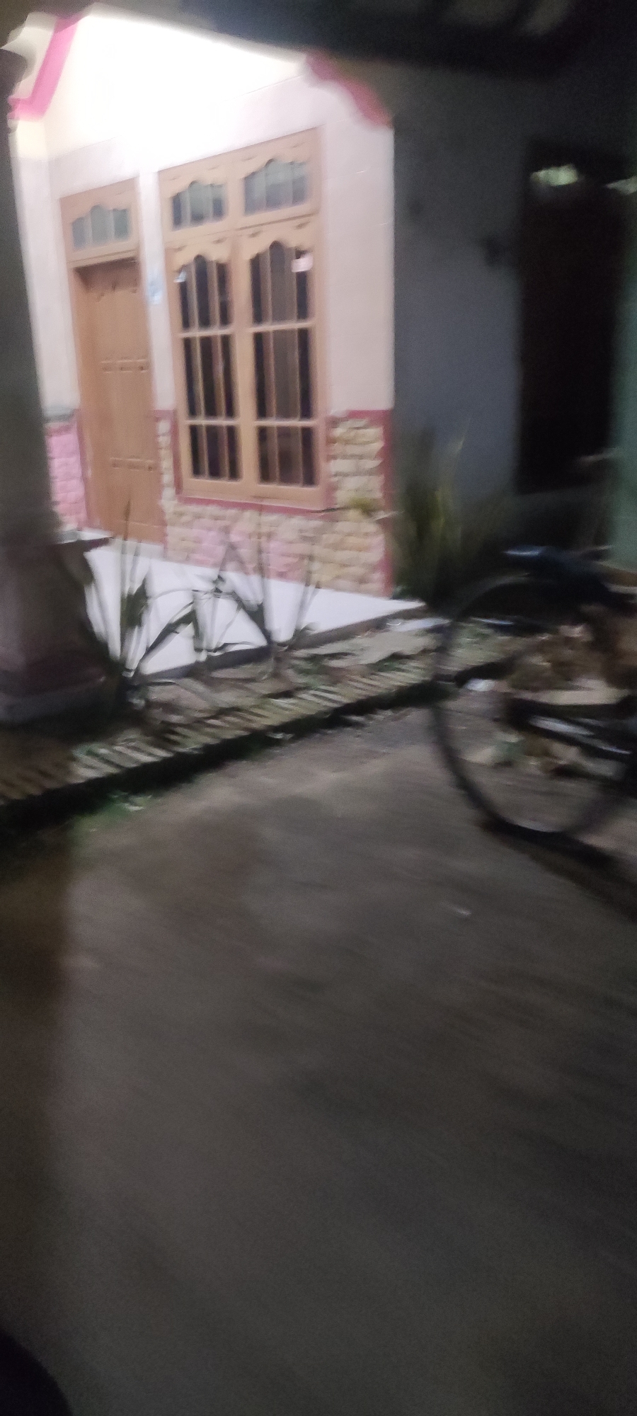 The image shows a house in Gebang, Indonesia, at night. The house is a single-story structure with a light-colored exterior and a prominent wooden window with multiple panes. To the left of the window is a wooden door. The lower part of the house's facade is adorned with a patterned brickwork. In front of the house, there's a tiled pathway leading up to the door and window. Several thin, green plants are situated along the pathway. On the right side of the image, a bicycle is visible, leaning against something, with its wheel prominently featured. The ground in front of the house appears to be wet, suggesting recent rain or moisture. The overall lighting suggests it is nighttime, with artificial lights illuminating the house.