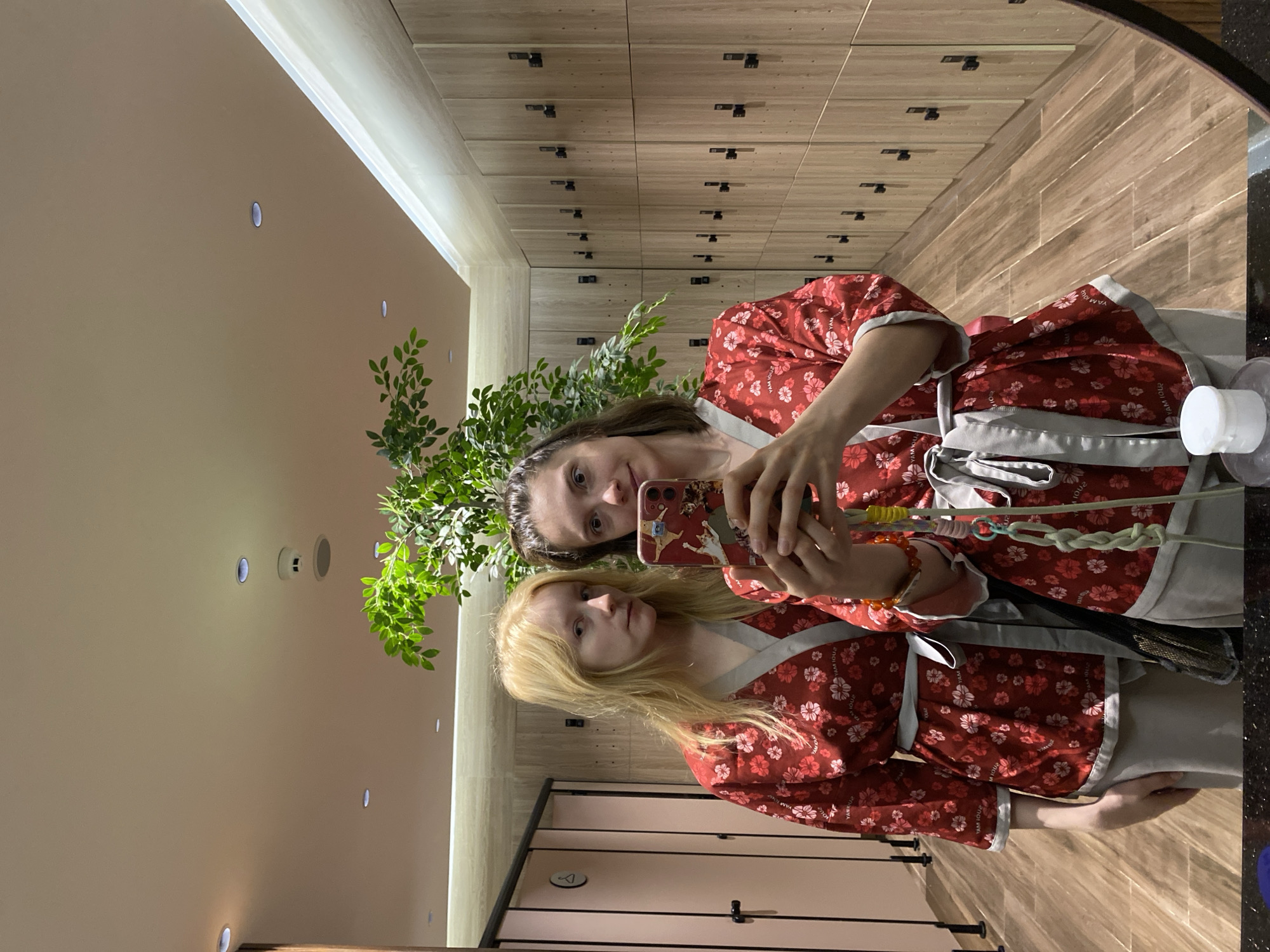 Two women are taking a mirror selfie in a locker room. They are both wearing red robes with a floral pattern and grey trim. The woman on the left has blonde hair and is looking at the camera with a neutral expression. The woman on the right has dark hair and is holding a phone up to take the photo, smiling slightly. The locker room has wooden lockers lining the wall and pink shower stalls visible on the left. A large potted plant with green leaves is positioned behind the women. The floor is made of wood planks. The ceiling has recessed lighting and a smoke detector. The overall lighting is warm and inviting. The image is taken from a slightly high angle. There is a white bottle of lotion visible in the bottom right corner of the mirror's reflection. There is no visible indication that this location is in Da Lat, Vietnam.