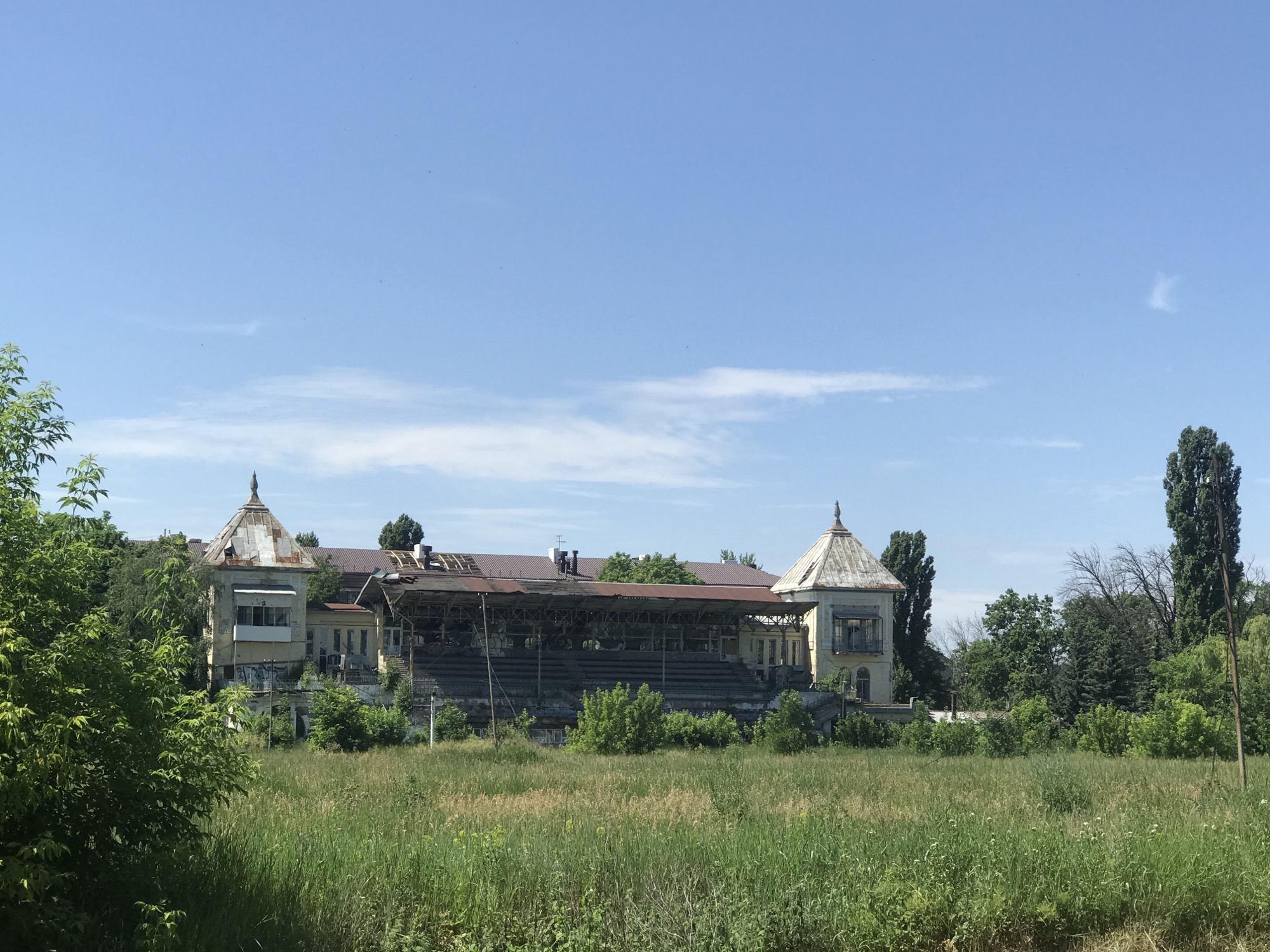 This is a horizontal, eye-level shot of an abandoned stadium in Kharkiv, Ukraine, under a bright blue sky with scattered clouds. In the foreground, tall green grass and weeds cover a field. Behind the field, the stadium structure dominates the midground. The stadium features weathered grandstands with tiered seating, partially covered by a rusty, decaying roof supported by metal beams. The building connected to the grandstands is yellow and shows signs of neglect, with graffiti visible on its walls and broken windows. Ornate, conical towers with weathered roofs top sections of the building. Lush green trees and bushes surround the stadium, with taller trees, including a very tall, slender one on the right, framing the scene. No people are visible.