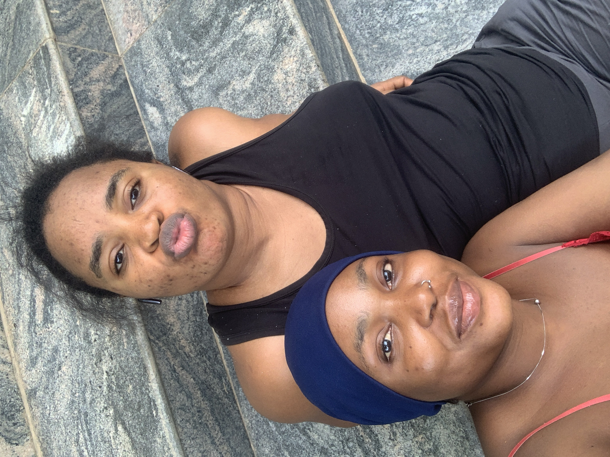 Two young women are photographed from a high angle, outdoors, with a textured grey stone surface as the background. The woman in the foreground, on the left, is wearing a blue head covering, a thin silver necklace with a small bead, and a red strap top. She is looking directly at the camera with a slight smile. The woman behind her, on the right, is wearing a black tank top and has her hair pulled back. She is also looking at the camera, with her lips puckered into a kiss. The scene appears to be a casual, everyday moment captured in Abuja, Nigeria.