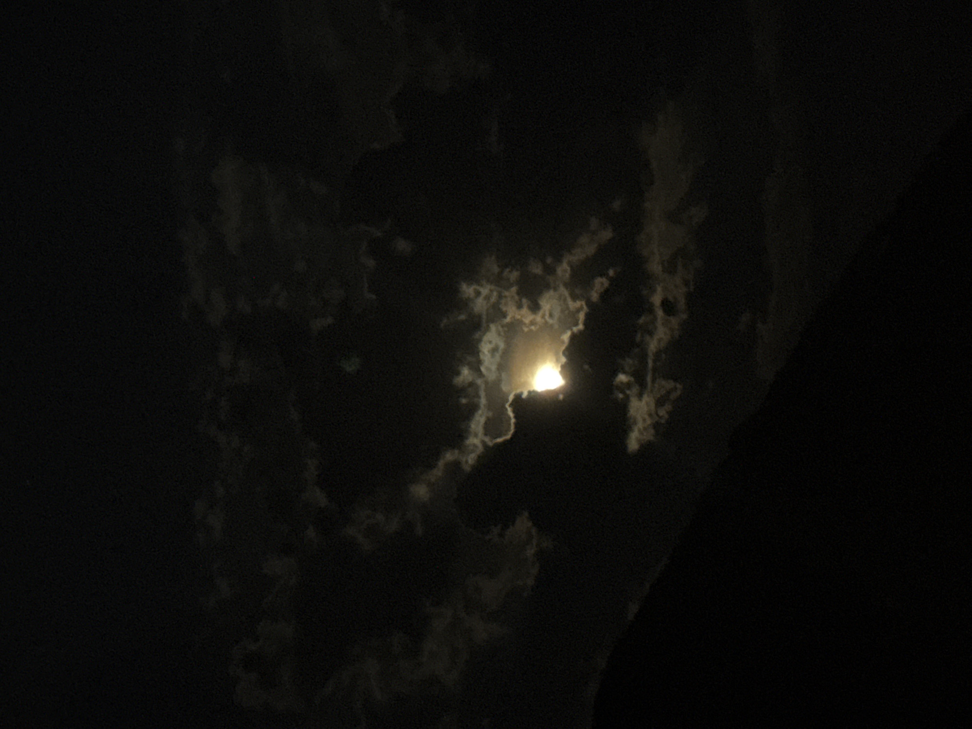 This nighttime image captures a dramatic view of the moon partially obscured by clouds, set against a dark sky, likely in Budva, Montenegro. The primary subject is the moon, which glows with a bright, yellowish-white light, emerging from behind thick, dark cloud formations.
The clouds vary in density and shape, with some appearing dense and opaque, blocking most of the moonlight, while others are thinner and more translucent, allowing the light to diffuse and create soft glows around their edges. The moon is positioned towards the upper-right of the frame, casting its light to illuminate the intricate textures and outlines of the surrounding clouds. The interplay of light and shadow creates a dynamic and somewhat mysterious atmosphere.
The overall scene is dark, emphasizing the brightness of the moon. The background is a deep, velvety black, indicating a clear night sky beyond the immediate cloud cover. On the far right edge of the image, a dark, indistinct silhouette runs vertically, possibly representing part of a landmass, a building, or tall trees, further framing the celestial display. There are no visible people, specific man-made objects within the illuminated area, or any discernible text. The image evokes a sense of quiet grandeur, highlighting the natural beauty of a moonlit, cloudy night.